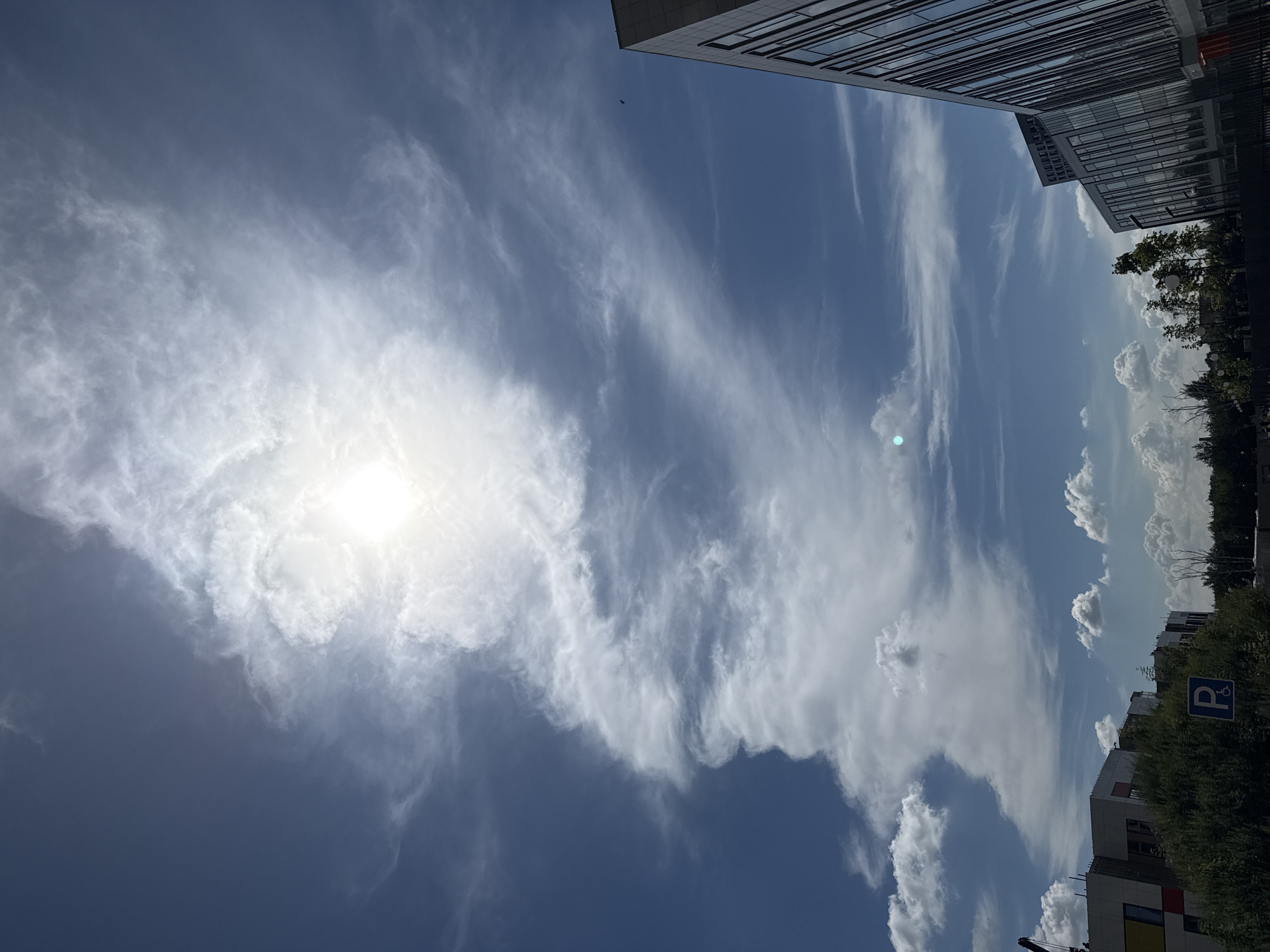 The image shows a daytime view of a modern urban environment in Lyubertsy, Russia. The sky is a dominant feature, filled with scattered white clouds against a deep blue background. The sun is bright and partially obscured by wispy clouds, creating a luminous effect in the upper center of the frame. In the foreground, on the right side, a contemporary building with a glass facade is visible, angled towards the viewer. On the left side, another building, possibly residential or commercial, with colorful sections is partially seen, along with some greenery. A parking sign with a wheelchair symbol is also visible among the trees. The overall scene suggests a clear, possibly sunny day in a developed area.