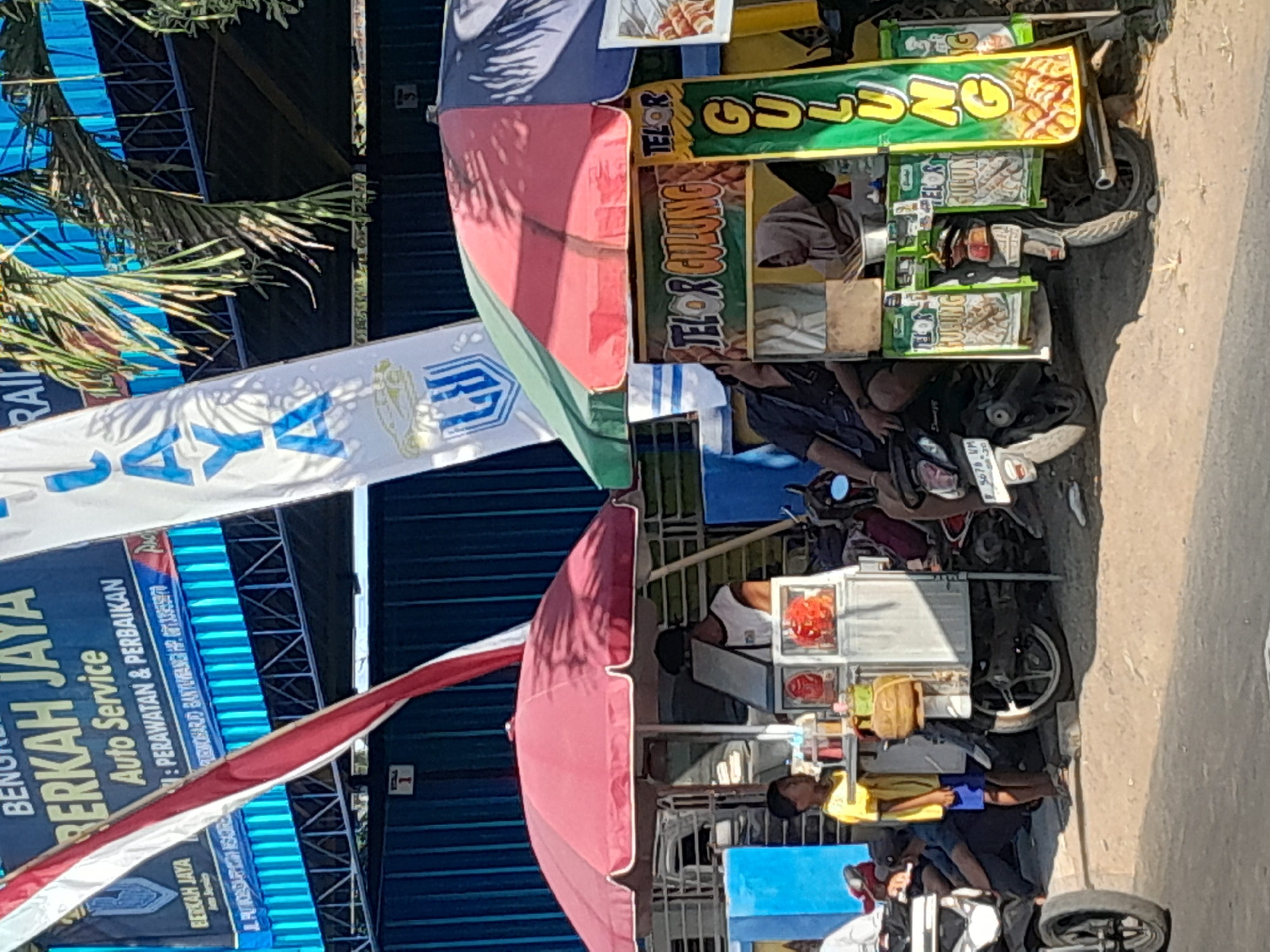 The image depicts an outdoor street scene in Bulurejo, Indonesia, under bright daylight. Two food carts are prominent. The cart on the left, covered by a red umbrella, displays food items and appears to be staffed by a person wearing a white shirt and a dark cap. A young boy in a yellow shirt and blue shorts stands to the left of this cart, looking away from the camera. The cart on the right, shaded by a multi-colored umbrella (green, red, and blue), advertises "TELOR GULUNG" (rolled eggs) with signage showing images of the food. A woman in a hijab is visible behind this cart, attending to the food. Several motorcycles are parked around the carts, with people seated on some of them. In the background, banners advertising "BERKAH JAYA AUTO SERVICE" and "RAIC" are visible, along with a blue structure that appears to be part of a building or fence. Palm trees cast shadows across the scene. The ground is unpaved, suggesting a dusty or sandy environment.