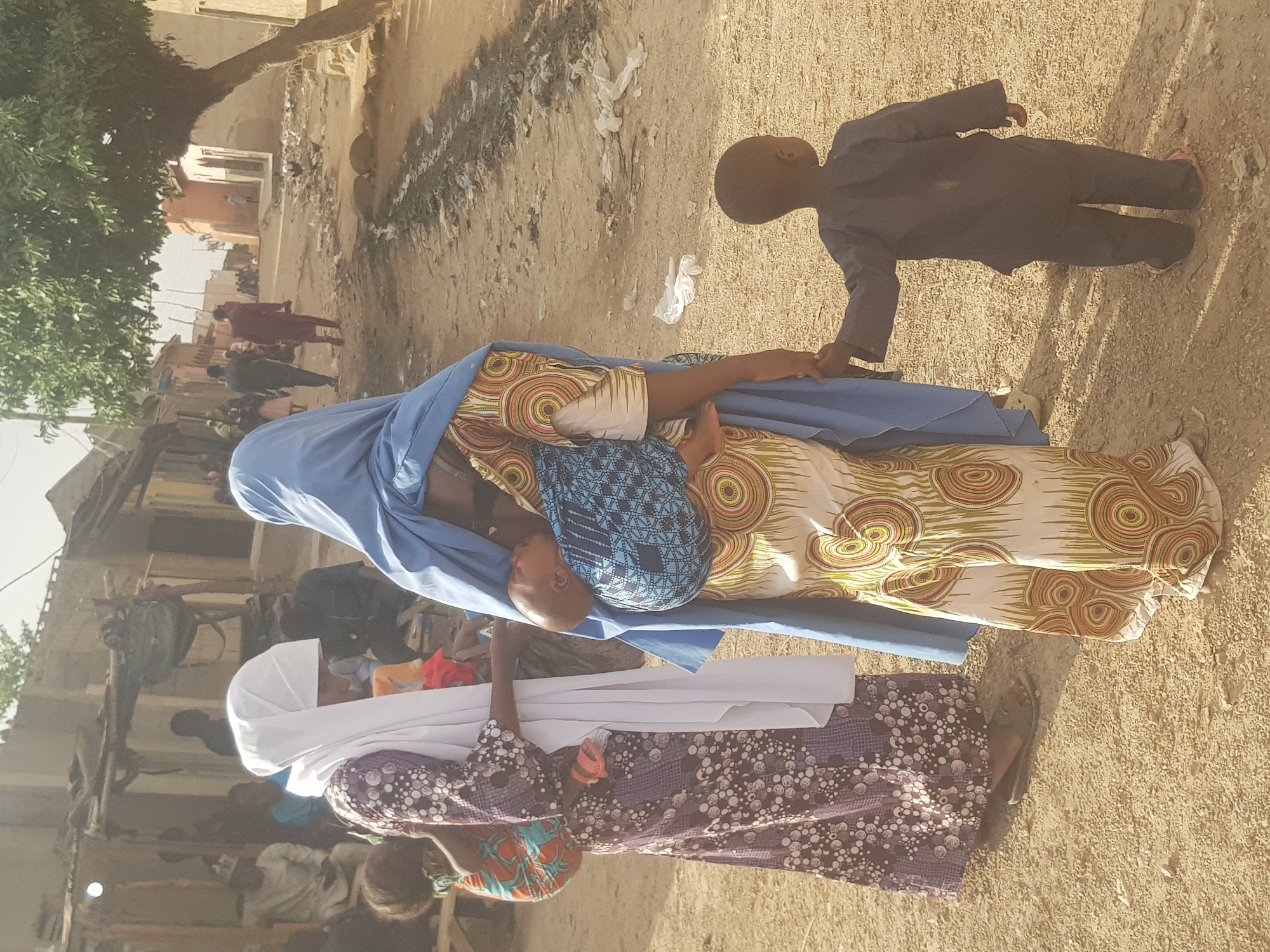 The image depicts a street scene in Dutsinma B, Nigeria. A woman wearing a patterned dress and blue hijab is breastfeeding an infant covered in blue fabric. A young boy stands beside her, holding her hand. Another woman wearing a patterned dress and white hijab is kneeling. The ground is dirt, and there is litter visible. In the background, there are buildings, people, and a tree.