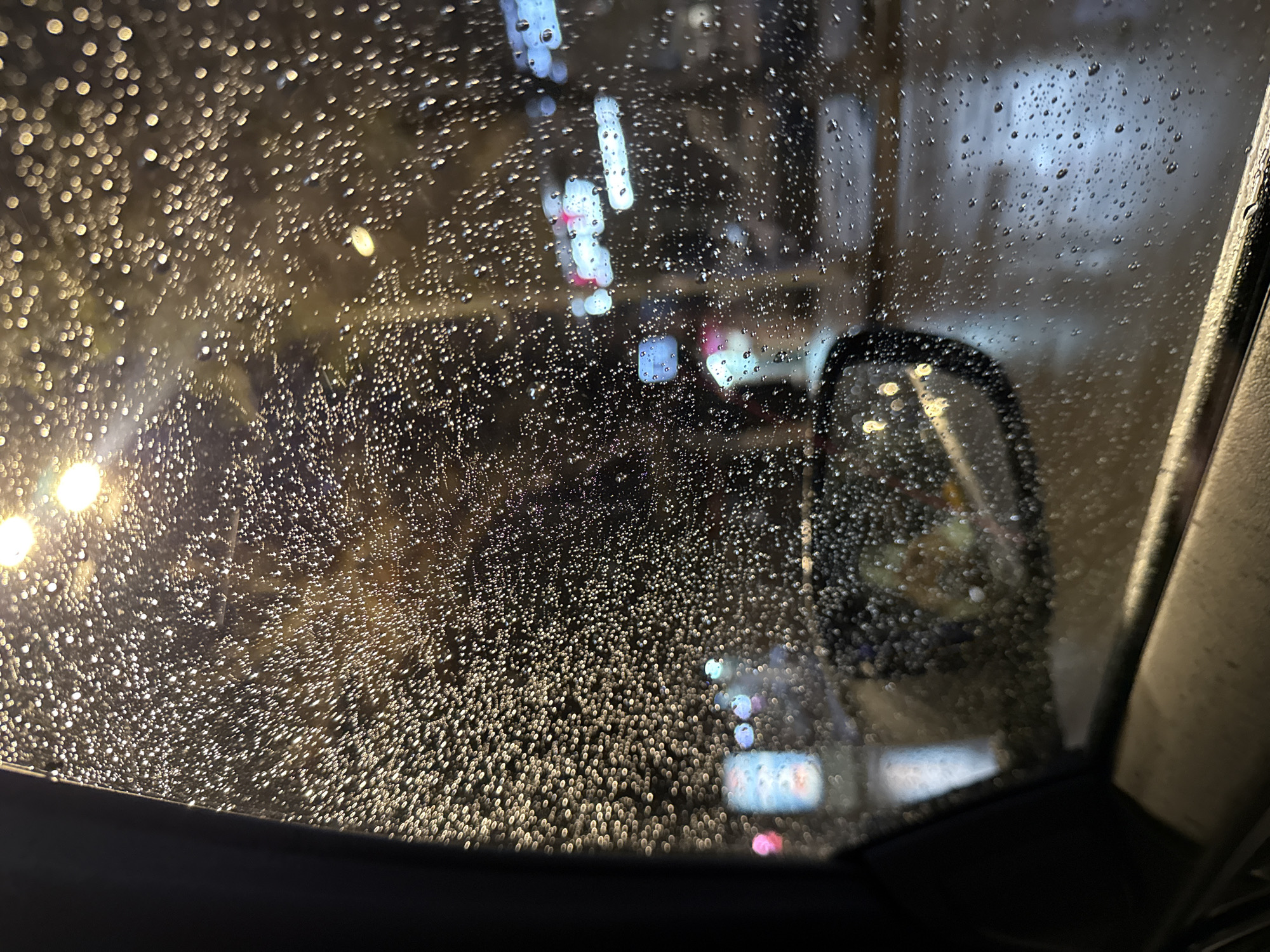 The image shows a view through a rain-covered car window at night. Outside, there are blurred streetlights and building lights suggesting an urban environment. A car side mirror is visible in the background. The location is not definitively identifiable as Yerevan, Armenia based solely on the image content. The droplets on the window obscure the view, creating a bokeh effect.