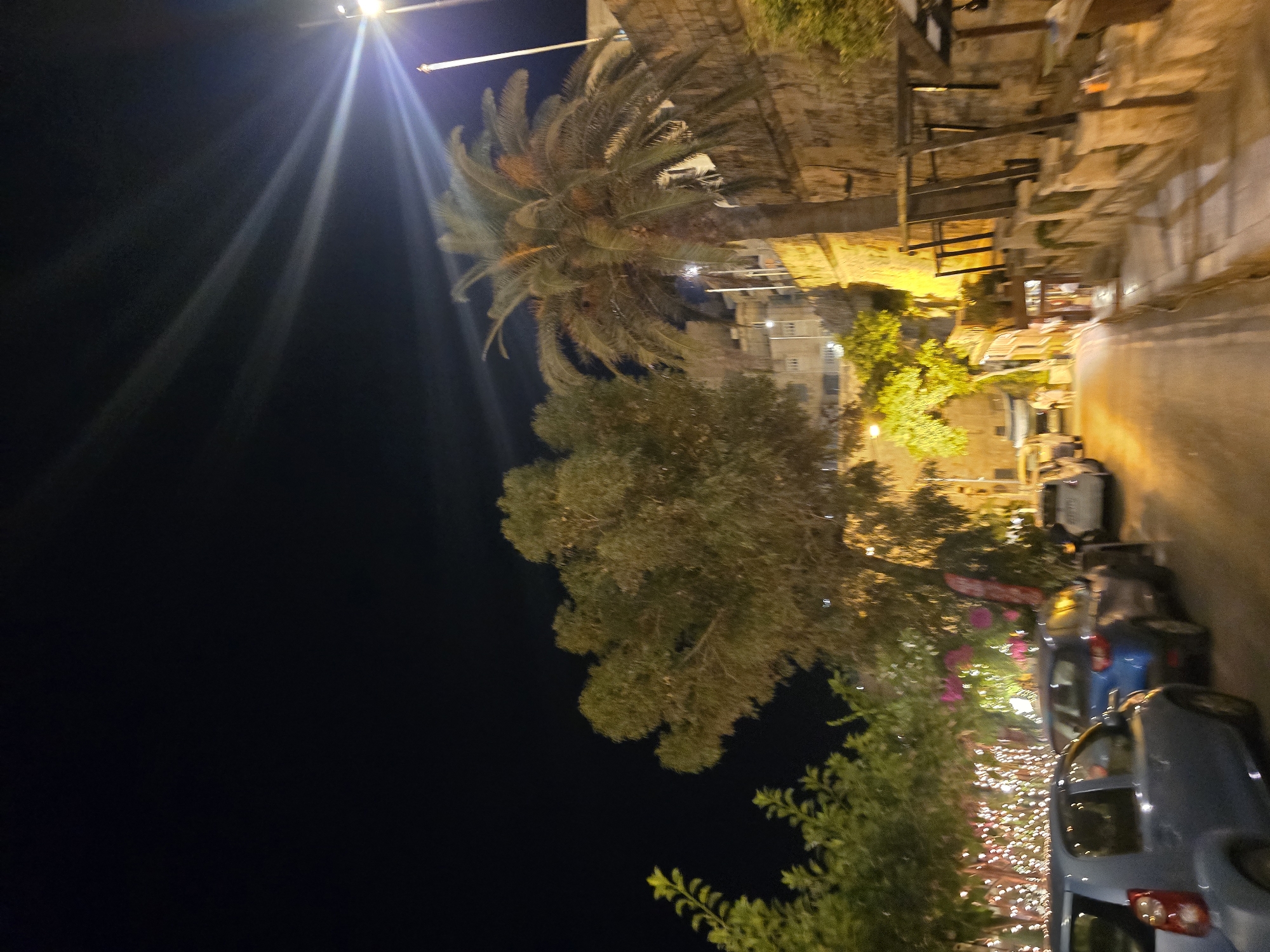 The image depicts a street scene at night in Valletta, Malta, with a focus on the illumination from streetlights and decorative lights. In the foreground, several cars are parked along the right side of the street, illuminated by the ambient light. A palm tree stands prominently on the right, with its fronds catching the light. To the left, a line of trees adorned with numerous small, bright lights creates a festive atmosphere.
In the background, stone buildings with illuminated windows suggest urban density. The street itself is paved and slopes gently upwards and to the right. The sky is dark, with bright beams of light radiating from a source not fully visible in the frame, likely a powerful spotlight or street lamp. The overall impression is of a well-lit, possibly tourist-oriented area in Valletta at night.