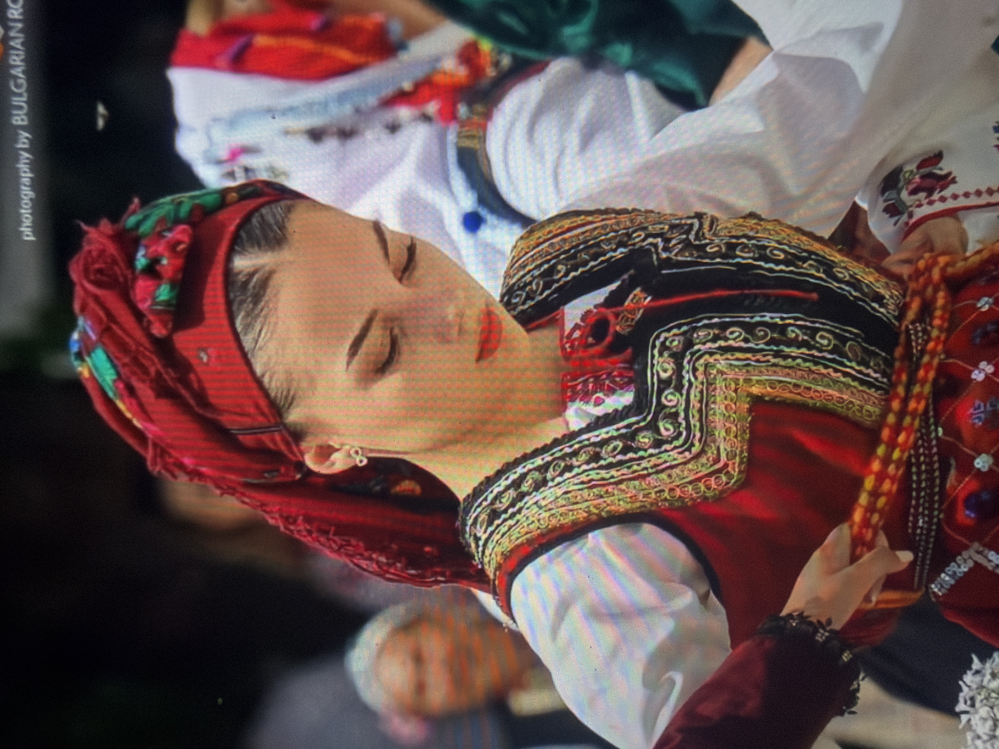 The image displays a young adult female, positioned slightly off-center to the right, engaged in a cultural performance or display within the city of Shumen, Bulgaria. She is captured looking downwards with a focused expression, her lips accentuated with red lipstick. Her dark hair is parted in the middle and neatly styled beneath a red headscarf, which is adorned with green, red, and blue floral embroidery along the front and top. A small, silver-colored earring shaped like an infinity symbol is visible in her right ear.
She is dressed in elaborate traditional attire. She wears a white, long-sleeved blouse with subtly puffed sleeves, over which a dark-colored (likely black or dark brown) sleeveless vest is worn. The vest is intricately embroidered with gold, black, and red swirling patterns. A red skirt or dress is visible beneath the vest and around her waist. Both hands are holding and adjusting a multi-strand beaded belt or sash, primarily composed of red and yellow/gold beads, positioned around her lower abdomen. The cuff of her left sleeve appears dark, possibly embroidered or made of a different material.
The background is out of focus, indicating a shallow depth of field. To the left, a blurred older individual with grey hair is partially visible. To the right, another individual in a white garment with red and blue traditional elements is also out of focus. The overall background is dark and indistinct, suggesting an event setting.
In the upper right corner of the image, partially visible white text reads "photography by BULGARIAN RO". The image exhibits visual artifacts consistent with a photograph of a screen, including noticeable pixelation, color fringing, and a subtle grid pattern across the entire scene.