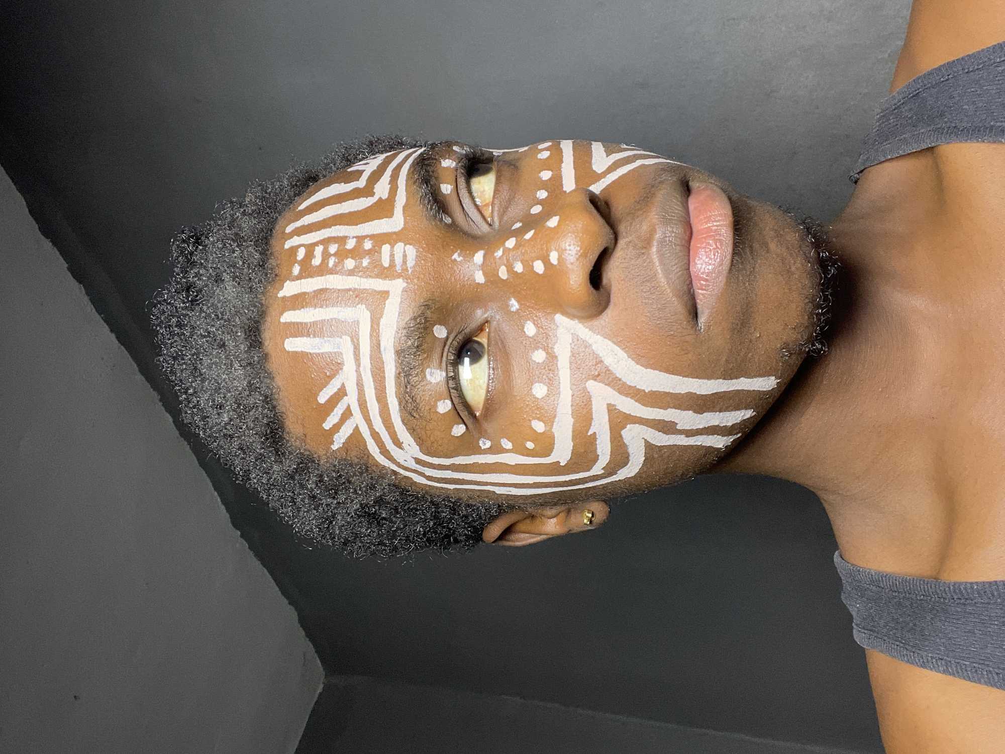 A medium close-up, low-angle shot shows a person with dark skin and short, dark curly hair, facing upwards. The person's face is adorned with white, painted tribal-like markings including lines and dots. The background is a two-toned gray. The person wears a gray top. The setting is not discernible as Nkpor, Nigeria.