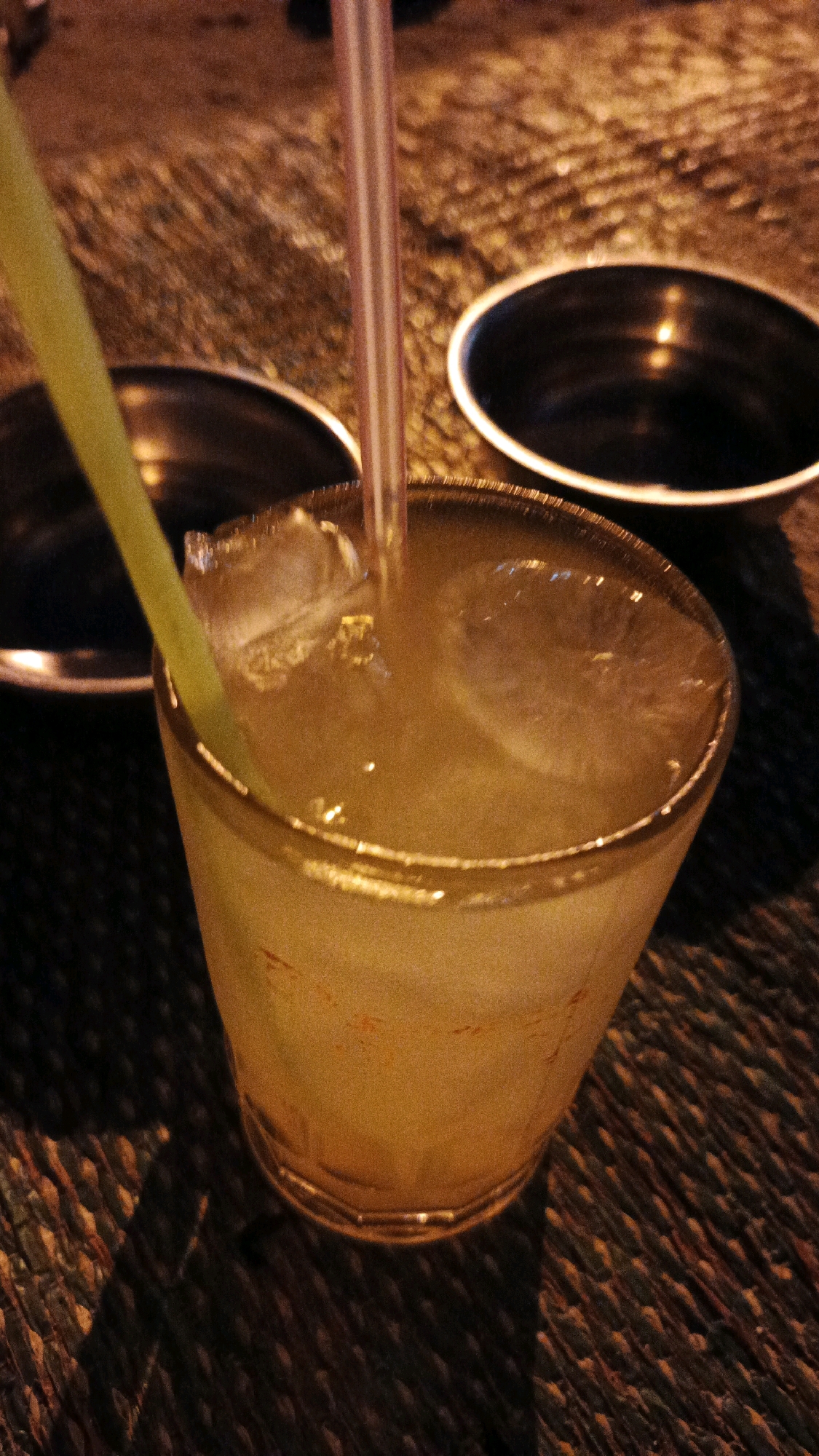 The image shows a close-up, low-angle shot of a glass of yellow beverage with ice and two straws, one yellow and one pink, in a dimly lit outdoor setting. Two dark, shallow bowls are visible in the background, one on the left and one on the right. The beverage is in a clear, slightly tapered glass. The surface on which the glass and bowls rest appears to be a woven mat or textured surface, possibly a table. The lighting suggests it is evening or night. The scene is likely at an outdoor food stall or cafe in Surakarta, Indonesia, given the commonality of such setups in that region. There are no people or identifiable human interactions depicted. The overall impression is one of a casual, relaxed setting, possibly a street-side eatery.