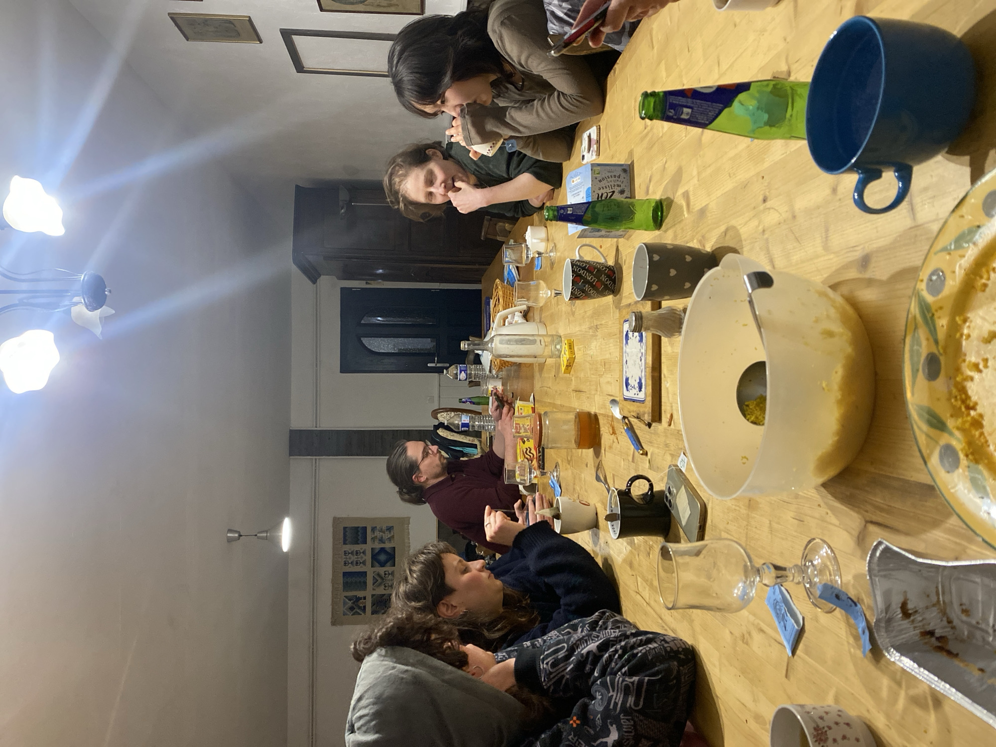 A group of approximately six people are gathered around a long wooden table indoors. The setting appears to be a dining room or communal space in Cornillon-en-Trièves, France, judging by the style of the architecture and furnishings. The table is laden with food and beverages, including bottles, mugs, plates, and a large mixing bowl containing what appears to be a batter or dough. Some individuals are looking at their phones, others are talking. Dishes and serving ware indicate that a meal has been eaten. The room is lit by a chandelier overhead and a wall sconce. Artwork and a patterned cloth hang on the walls. A partially-opened dark door leads to another room.