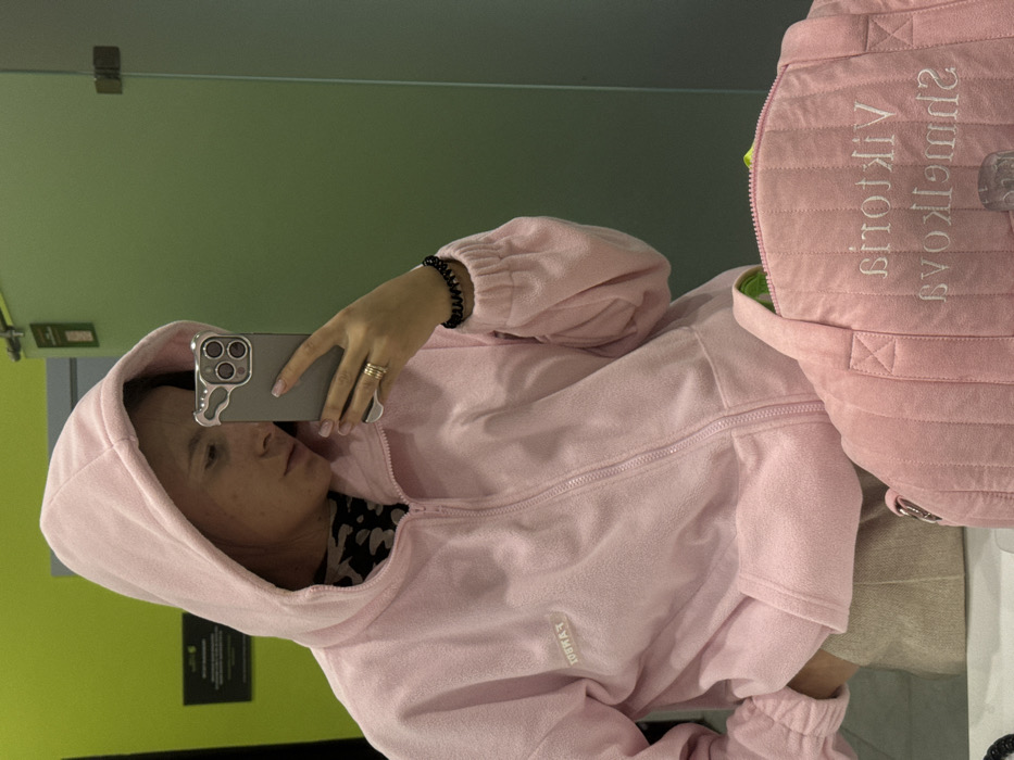 The scene depicts a young adult female taking a mirror selfie in an indoor setting, located in Vniissok, Russia. The individual, seen from the chest up, wears a light pink fleece hooded jacket with a front zipper and a white rectangular patch bearing the word "FANBOX" on the left chest. A dark, possibly leopard-print, top is visible beneath the jacket, and light beige or off-white pants are seen below the jacket's hem. She holds a gray smartphone, identifiable as an iPhone Pro model with a silver-colored, irregularly shaped case, in her left hand, directing it towards the mirror. Gold rings adorn her left ring finger, and a black spiral hair tie is worn on her right wrist. Her fingernails are manicured with a light polish. In the lower right foreground, a pink quilted bag with two top handles rests on a surface. The bag displays white embroidered text, mirrored, which reads "Viktorija Smelkova." A glimpse of a bright green lining or item is visible within the bag's open zipper. The background features a wall divided into two sections: a bright lime green on the left and a muted sage green on the right. A dark green door with visible silver hinges is present on the right side of the frame. A dark rectangular sign is mounted on the upper left lime green wall.