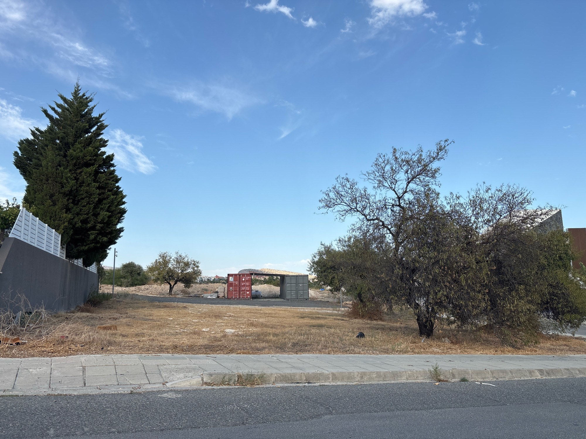A serene, open lot in Agios Athanasios Municipality, Cyprus, is depicted under a clear blue sky dotted with wispy white clouds. The foreground features a gray asphalt road adjacent to a concrete sidewalk with a curb. Beyond the sidewalk, the lot is covered in dry, yellowed grass and scattered debris, including a red brick and some plastic litter. A large, dark gray retaining wall runs along the left side of the lot, topped by a white fence. A tall, dark green cypress tree stands sentinel near the wall.
Further into the lot, a cluster of three shipping containers are visible. Two are red, and one is green, positioned next to each other. Behind them, a simple open-sided structure with a light-colored roof has been erected. Several smaller trees with sparse foliage are scattered across the landscape. In the distance, to the right, a modern building with a stone facade is partially visible. The overall impression is one of an undeveloped or transitional space within a municipality.