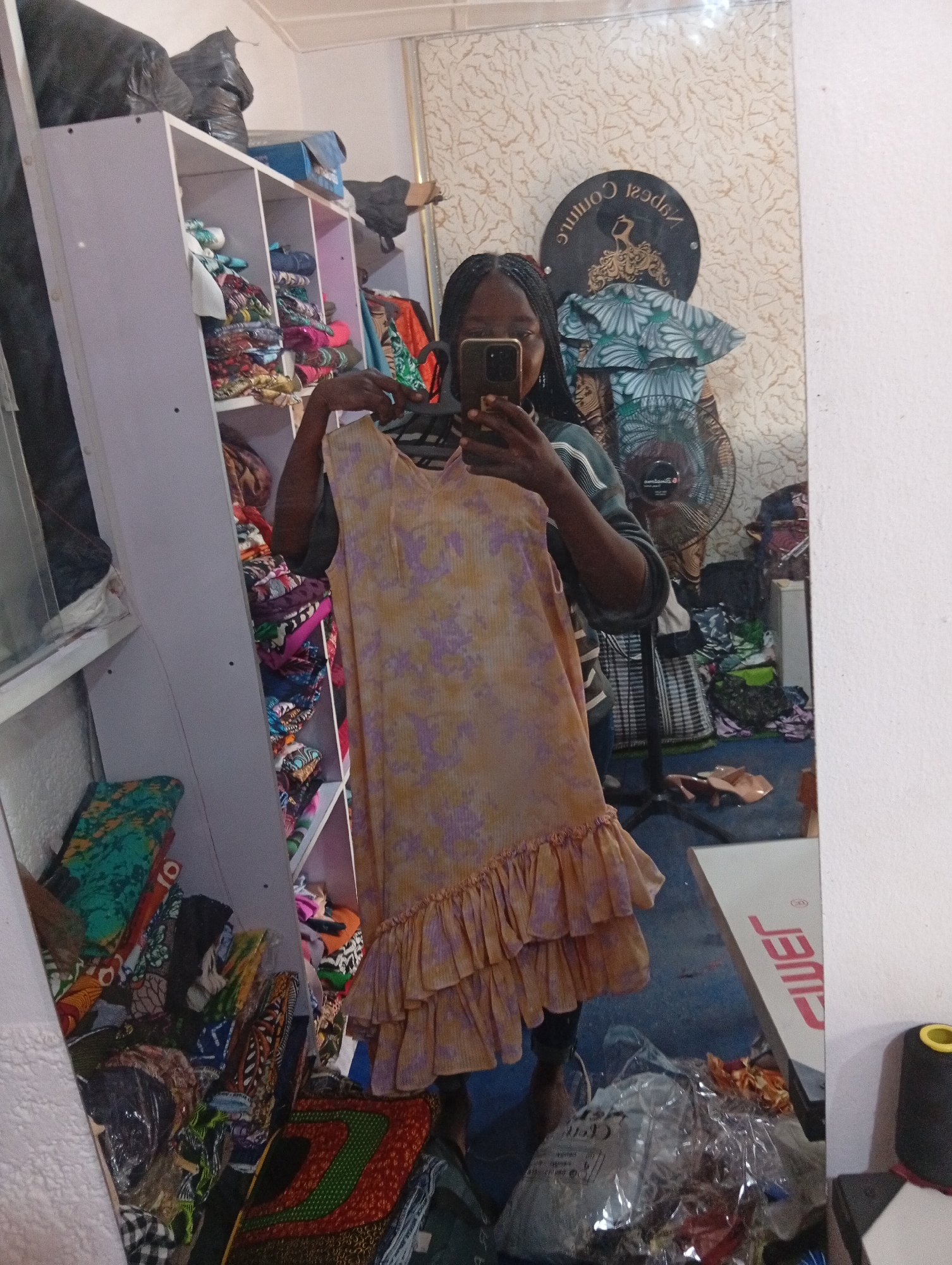 A person stands in front of a mirror, taking a selfie. The person is wearing a light-colored dress with a floral pattern and ruffled hem. They have dark braided hair.
The setting appears to be a room filled with fabric and sewing supplies, likely a tailor's shop or a seamstress's workspace. To the left, shelves are packed with colorful folded fabrics. In the background, visible in the mirror's reflection, is a large circular sign with text, a fan, and more stacks of materials.
The floor is a dark blue, and the walls have a textured wallpaper. A white table with red lettering is visible on the right side of the mirror. The overall impression is a cluttered but functional workspace. No specific time of day or weather conditions are discernible. The emotion conveyed is neutral, focused on the task of taking a photograph.