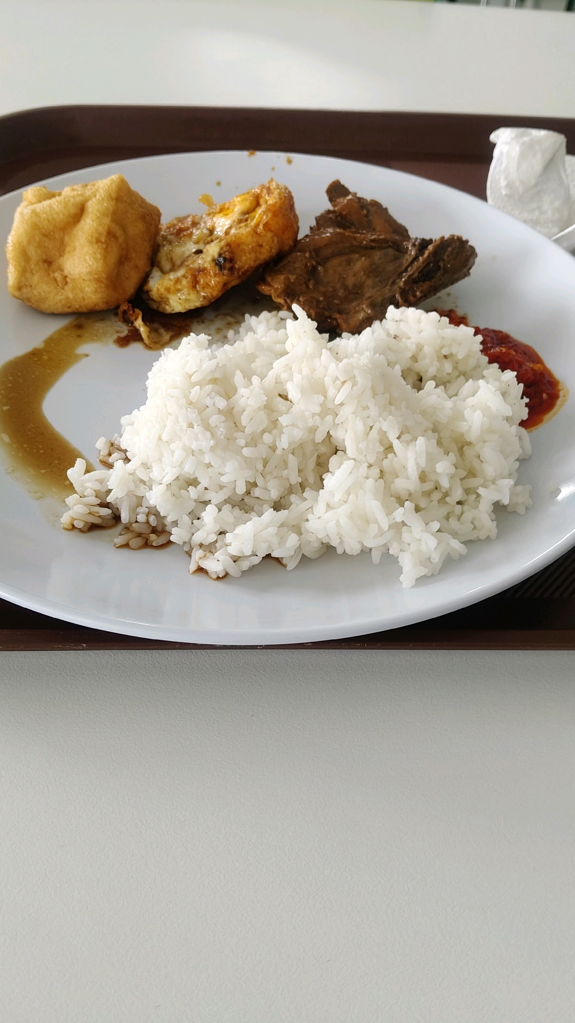 A close-up, high-angle shot shows a white plate of food, likely served in Mekarmukti, Indonesia. The plate is filled with a generous portion of white rice, piled high in the center. To the left of the rice are two pieces of fried food, one appearing to be a golden-brown puff of fried dough and the other a flatter, rectangular piece that looks like fried tofu or egg, with some filling visible. To the right of the rice, there is a piece of dark brown meat, possibly chicken or beef, in a sauce. A smear of red sauce, likely sambal, is visible on the far right edge of the plate. The plate rests on a dark brown tray, which in turn is on a white tabletop. A white napkin is crumpled in the upper right corner of the image. The background is out of focus, but appears to be a bright, neutral environment, consistent with a cafeteria or restaurant setting in Mekarmukti, Indonesia.
