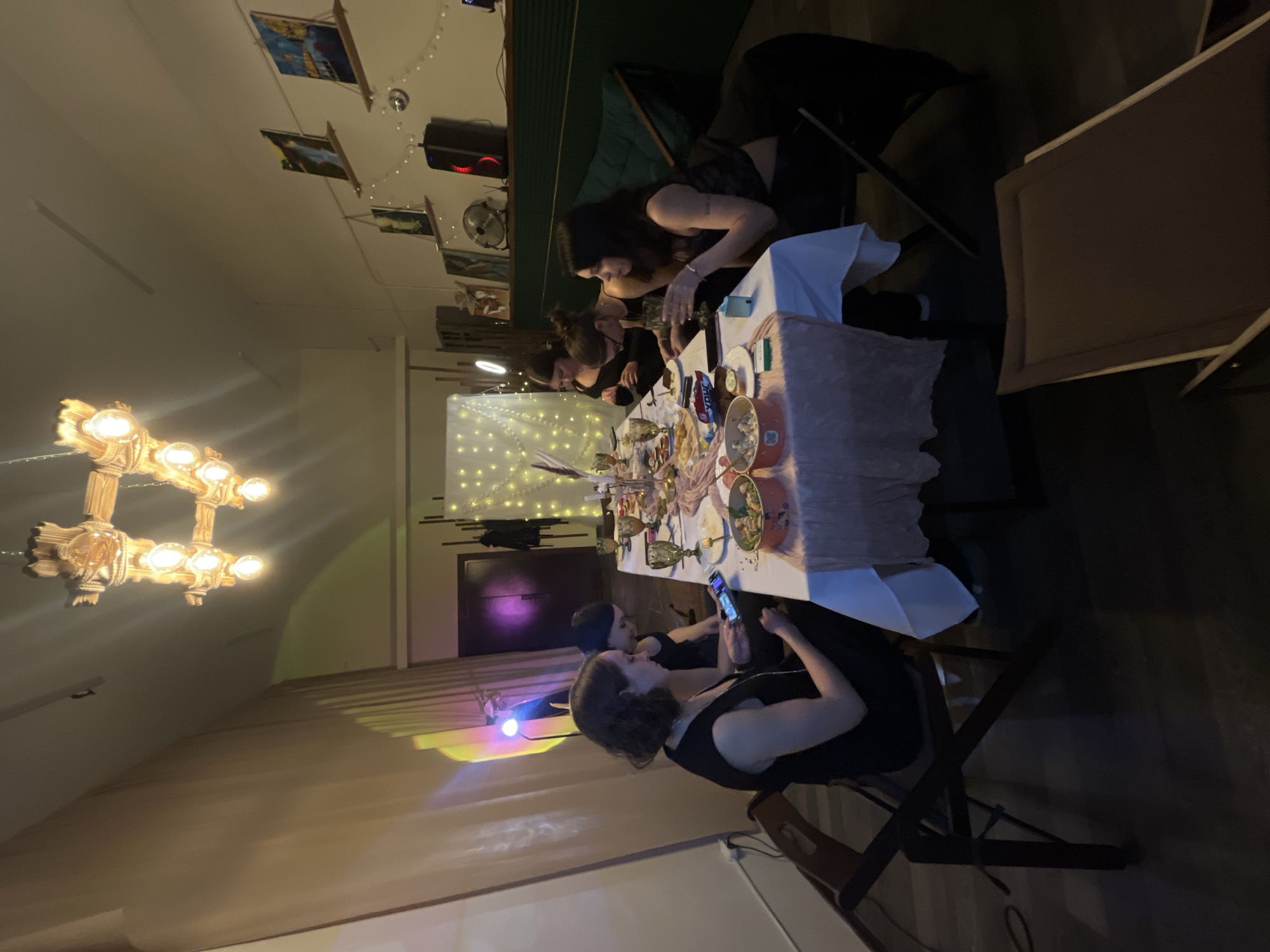 Here is a description of the image:
The image shows an indoor gathering, possibly a dinner or party, taking place in a room with somewhat dim lighting. A chandelier is visible on the left side. Several people are seated around a long table that is covered with a white tablecloth and various food items and glasses. Some individuals are looking at their mobile phones. There is a lighted backdrop, possibly a curtain or screen, with what appears to be string lights. Pictures are shown in the background. The location in the city of Saint Petersburg, Russia cannot be determined from the image.