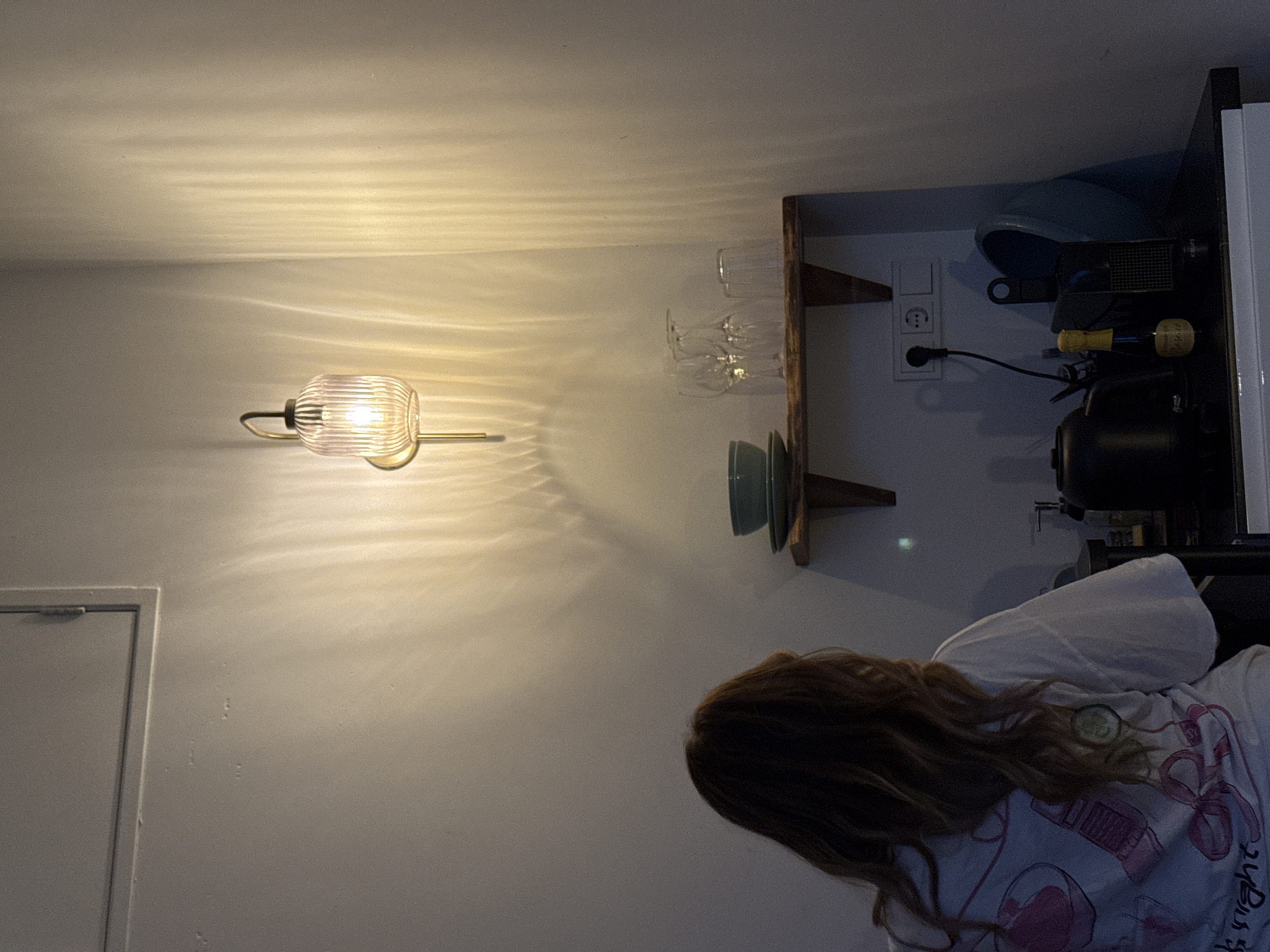 A low-angle, indoor shot displays a room with a person visible from the back, wearing a white t-shirt with a pink design. The person is looking toward a shelf that is mounted on the wall. The shelf is holding glassware and plates. Adjacent to the shelf is an electrical outlet with a cord plugged in. To the left of the shelf, a wall lamp is affixed to the wall, casting a patterned light. To the left of the lamp, there is a closed door. The image was taken indoors at an unknown location. There is no evidence suggesting that the location of the image is in Amsterdam, Netherlands.