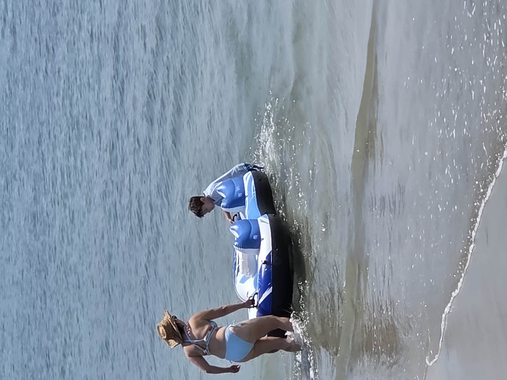 This media file shows two people near an inflatable watercraft on a beach. The scene is set in Long Beach, United States of America. The water is a calm, blue, and the waves are gentle, suggesting pleasant weather and possibly mid-day.
The first subject is a woman in a light blue and white bikini, wearing a straw hat. She is standing in the shallow water, with her back mostly to the camera. Her right arm is extended, and she appears to be holding onto the inflatable.
The second subject is a young person, likely a boy, wearing a white t-shirt. He is sitting on the inflatable watercraft, looking down towards the woman. The inflatable is predominantly white and blue, with black accents. It appears to be partially submerged in the water near the shore. The water is splashing around the inflatable, indicating movement.
The overall impression is a casual, recreational moment at the beach, possibly during a summer day. The lighting suggests it is daytime, with the sun casting a bright light. There are no visible text elements in the media file.