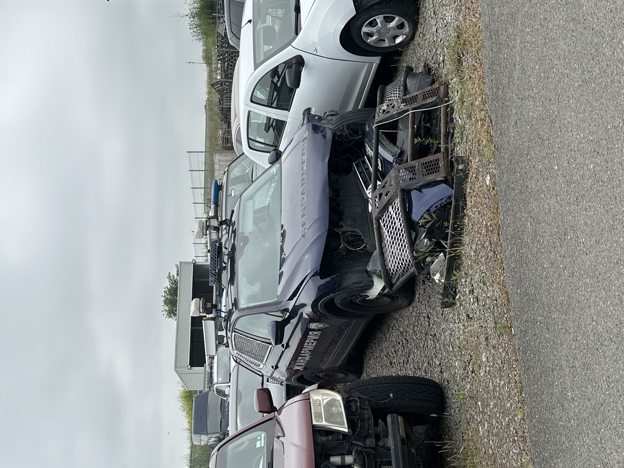 The image captures an outdoor vehicle storage or impound lot under an overcast sky. Multiple cars, many in a state of disrepair or with significant damage, are parked on a gravel surface adjacent to an asphalt road.
A prominent dark blue vehicle, appearing to be a law enforcement type, occupies the mid-ground. Its front end is heavily damaged, with the bumper and grille missing, exposing internal engine components. The side of this vehicle clearly displays the white Cyrillic text "ЖАНДАРМЕРИЯ" (Gendarmerie), indicating its function. It also features protective grilles on its windows and a roof rack or light bar assembly.
Behind the blue vehicle, several white passenger cars are visible, some also exhibiting damage. In the foreground, a maroon/reddish vehicle with a damaged front end is partially visible. The ground consists of light-colored gravel where the vehicles are parked, transitioning to a darker asphalt road. Sparse green vegetation grows along the edge of the gravel. In the background, there are fences, stacks of wooden pallets, and distant buildings, possibly industrial or storage facilities. The sky is uniformly overcast and bright, suggesting daytime conditions. No individuals are present in the scene.