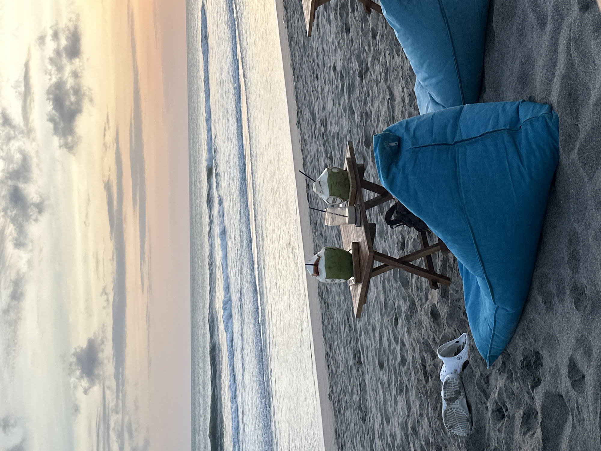 A tranquil beach scene at dusk in North Kuta, Indonesia, unfolds with soft, pastel-colored clouds gracing the sky. The gentle waves of the ocean create a rhythmic pattern on the shore, leading to a sandy beach where two low wooden tables are set up. On one table, two whole coconuts with straws are positioned next to glasses, suggesting a refreshing break. Beside these, a pair of white Crocs are casually placed on the sand, hinting at a relaxed atmosphere. The foreground is dominated by two large, plush blue beanbag chairs, inviting viewers to sit and enjoy the serene view. One beanbag chair is closer to the viewer, revealing its textured surface and a zipper. The other beanbag chair is partially visible to the right. A dark backpack is nestled on the sand between the tables. The overall ambiance is one of peaceful relaxation by the sea.