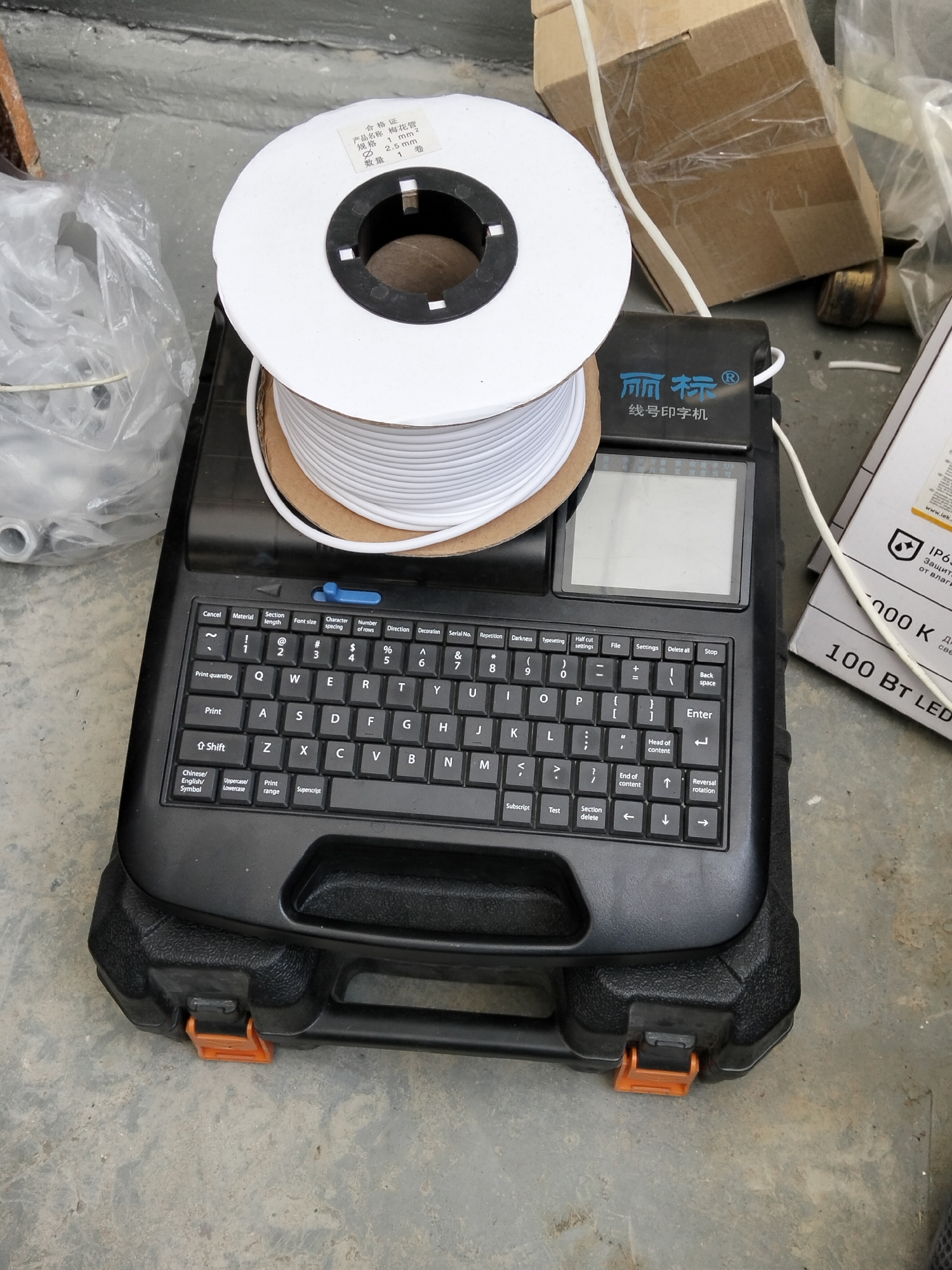 The image shows a black wire marker printer with a spool of white wire on top. The printer has a small screen and a QWERTY keyboard. The surrounding area appears to be an outdoor or industrial setting, possibly a construction site, with concrete ground and some cardboard boxes and plastic bags in the background. There are no people visible in the image. The spool of wire has a label with Chinese text, indicating "Product Name: Plume Pipe", "Specification: 1 mm² 2.5 mm", and "Quantity: 1 roll". The printer itself also has Chinese text, "丽标Ⓡ 线号印字机", which translates to "LibiaoⓇ Wire Number Printer". The overall impression is one of industrial equipment ready for use. The lighting suggests it might be daytime.