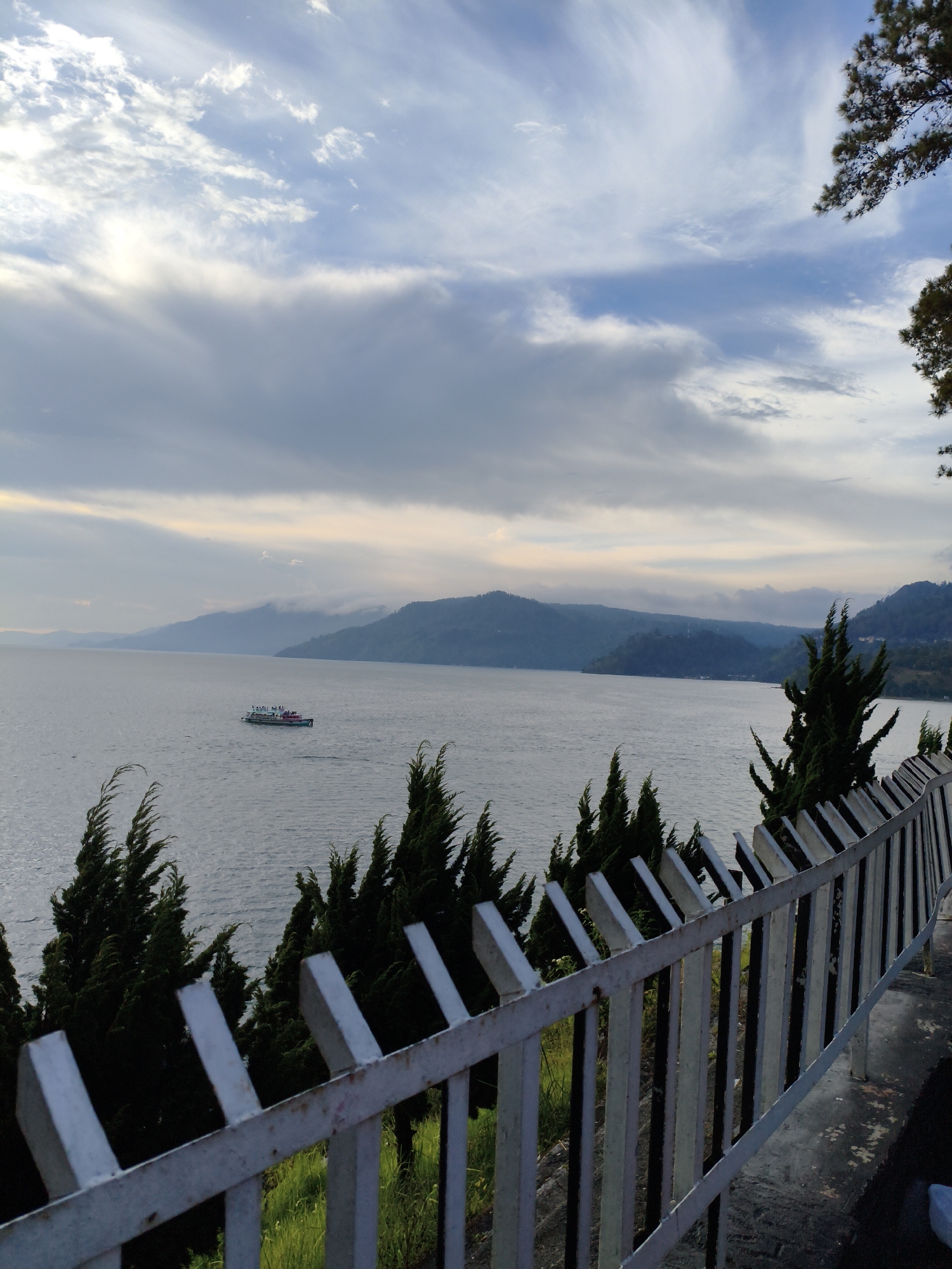 The image captures a scenic view of Lake Toba in Tiga Raja, Indonesia. In the foreground, a white metal fence runs diagonally across the frame, with the dark green foliage of coniferous trees visible behind it. The lake itself dominates the middle ground, its surface rippled by a gentle breeze. A white passenger ferry with a green stripe is visible on the water, carrying several people. In the background, rolling hills and mountains are shrouded in a soft, hazy light, with a cloudy sky overhead. The overall impression is one of tranquility and natural beauty, with the elements of human activity—the ferry and the fence—subtly integrated into the landscape.