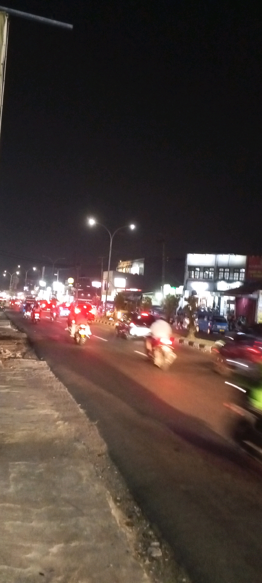 This is a nighttime, slightly motion-blurred, vertical-format image of a busy street in Cibinong, Indonesia. The foreground shows a concrete sidewalk bordering a paved road filled with traffic. Motorcyclists, cars, and possibly other vehicles are present, illuminated by streetlights and their own headlights and taillights, creating streaks of light due to the motion blur. The buildings lining the road are visible in the background, some with lit windows and signs. The overall impression is of a lively urban environment at night, with ongoing vehicular activity.