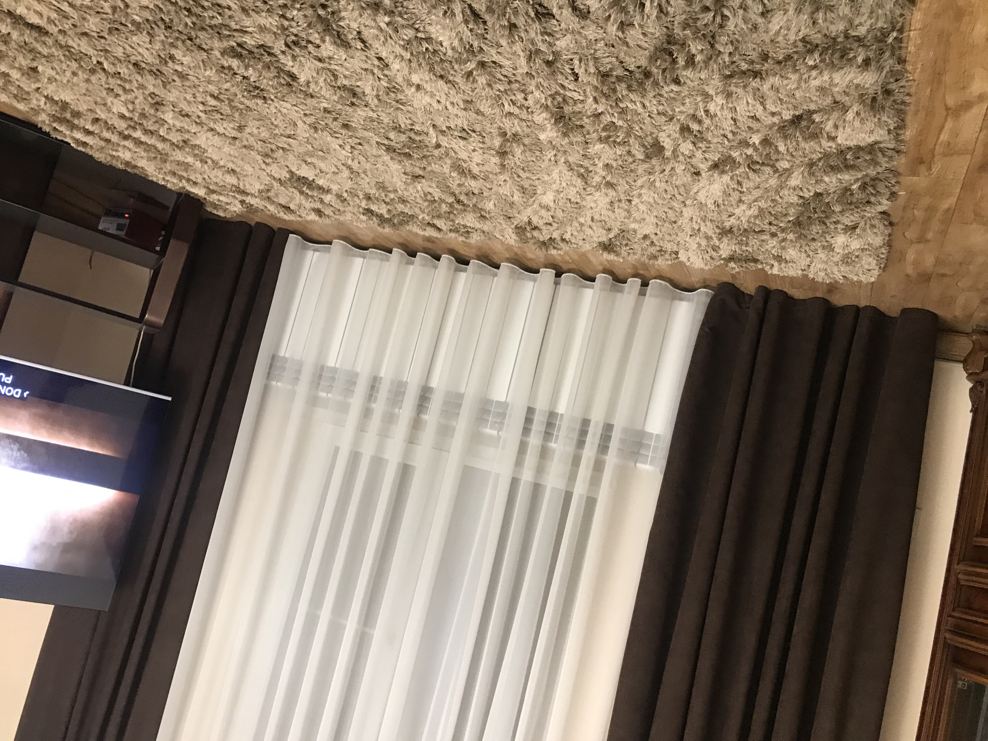 The image shows a room interior featuring a television screen on the left, framed by brown curtains. In the center, white sheer curtains filter light from a window. A shag rug covers the ceiling. The floor is wooden. The location is not discernable from the image. No people are visible.