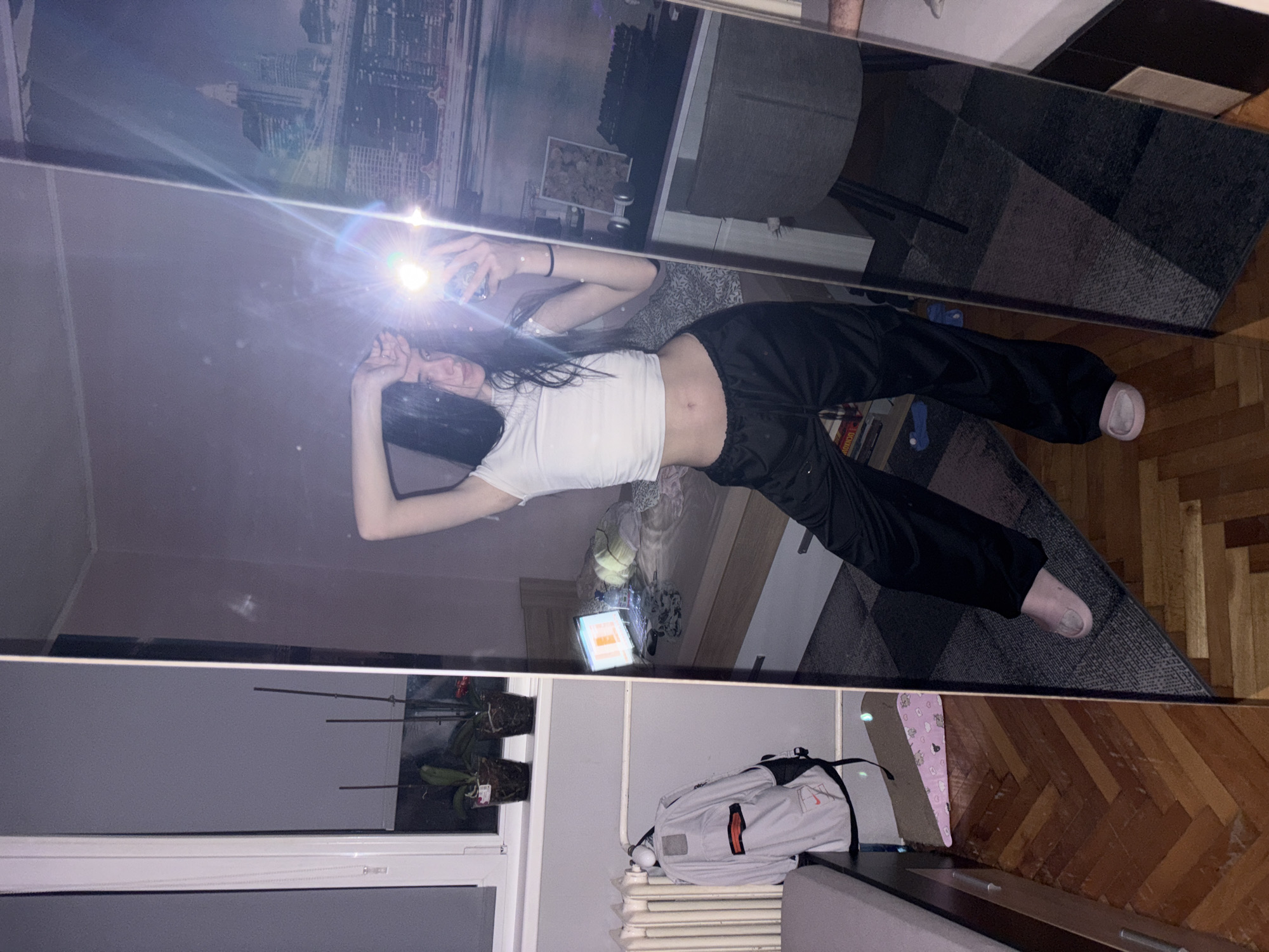 A young woman with long dark hair is reflected in a mirror as she takes a selfie. She is wearing a white crop top and black sweatpants. She is standing in a room with a window, radiator, and a backpack. The reflection in the mirror shows a cityscape, a bed, and a carpet. There is no visual data to determine if the location is in Sofia, Bulgaria.