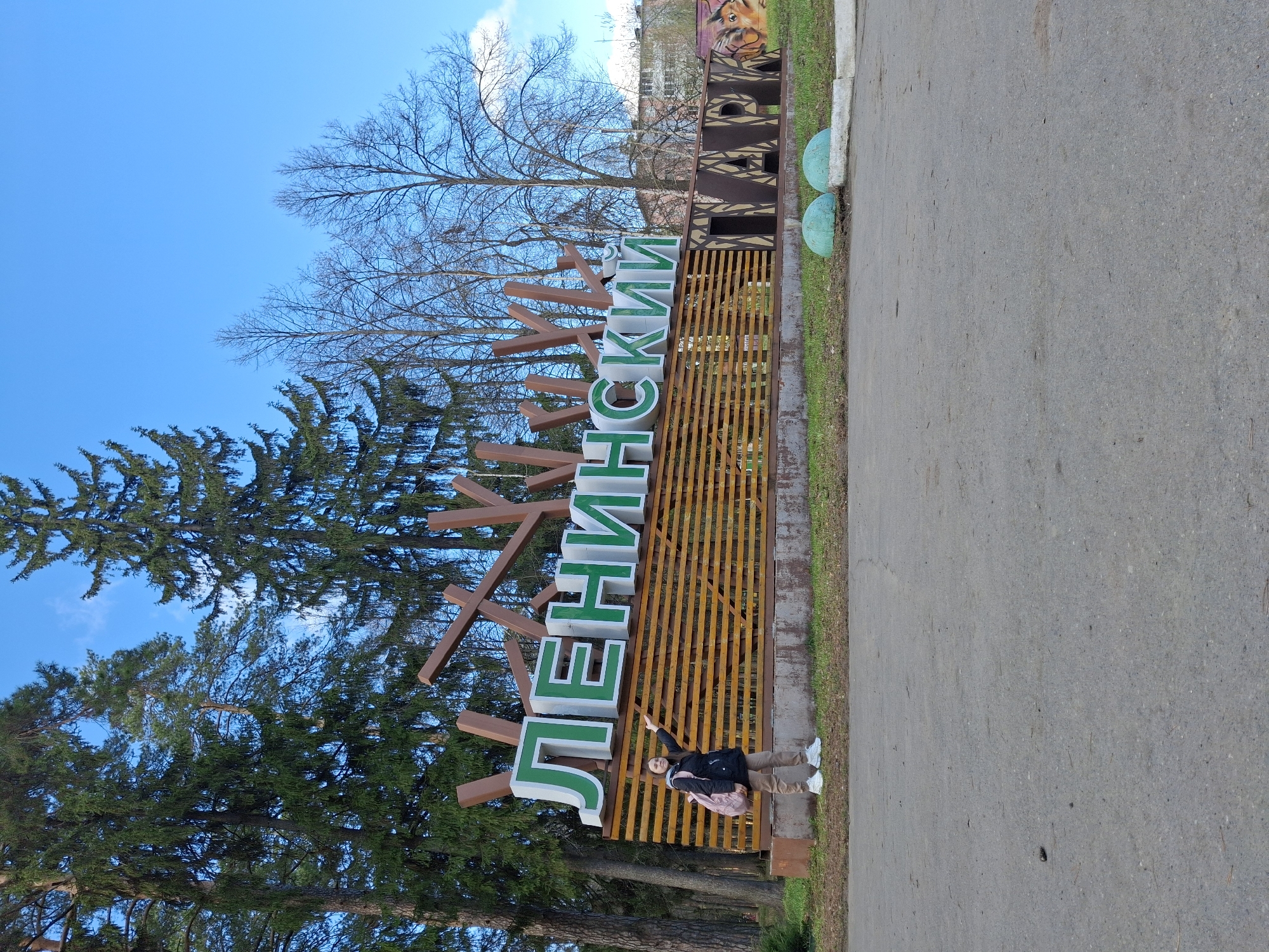 A daytime, tilted, medium shot shows a girl standing in front of the "Ленинский" park sign in the city of Sarapul, Russia. The green and brown sign sits on a brown, slatted, waist-high wooden structure. The girl is wearing a dark-colored jacket, light brown pants, and white shoes. She has her arm raised and is pointing at the first letter of the Russian word. She is wearing a pink backpack. To the left of the sign are tall green trees and a clear blue sky with some white clouds. To the right of the sign is a gray pavement path with grass and some greenery on the edge. In the background is a building and another sign with a picture of a lion. The light is diffused.