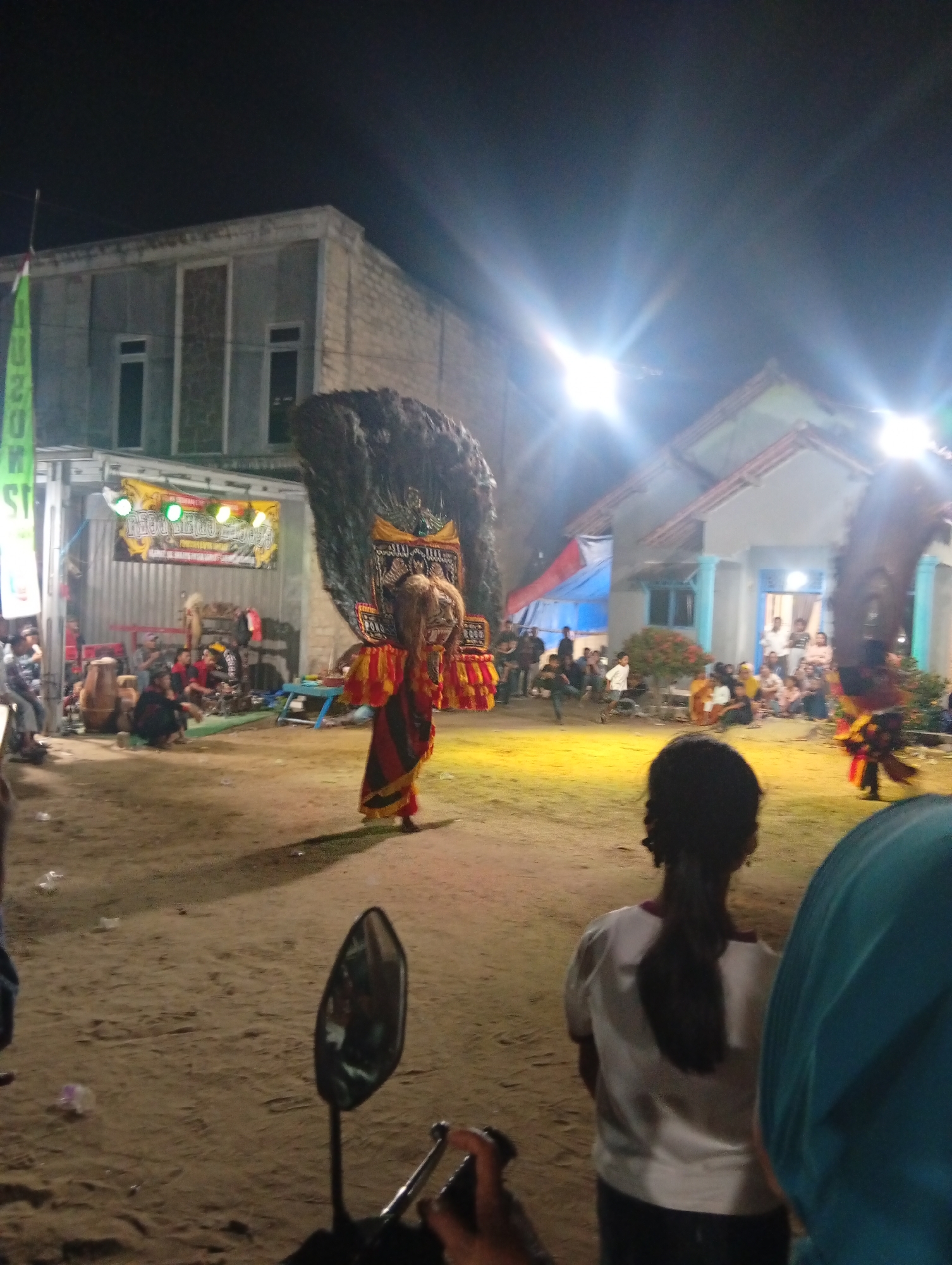 A nighttime outdoor performance is taking place in the city of Sri Gading, Indonesia. In the center of the frame, a performer wearing a large, elaborate costume with a prominent feathered headdress is dancing. The costume is red and yellow with intricate patterns. To the right, another performer in a similar costume can be seen. A crowd of spectators is gathered, some sitting and others standing, watching the performance. In the foreground, the back of a person's head with dark hair tied in a ponytail is visible, as is a motorcycle mirror and handlebars. The performance appears to be on a sandy, outdoor stage illuminated by bright stage lights. Buildings are visible in the background.