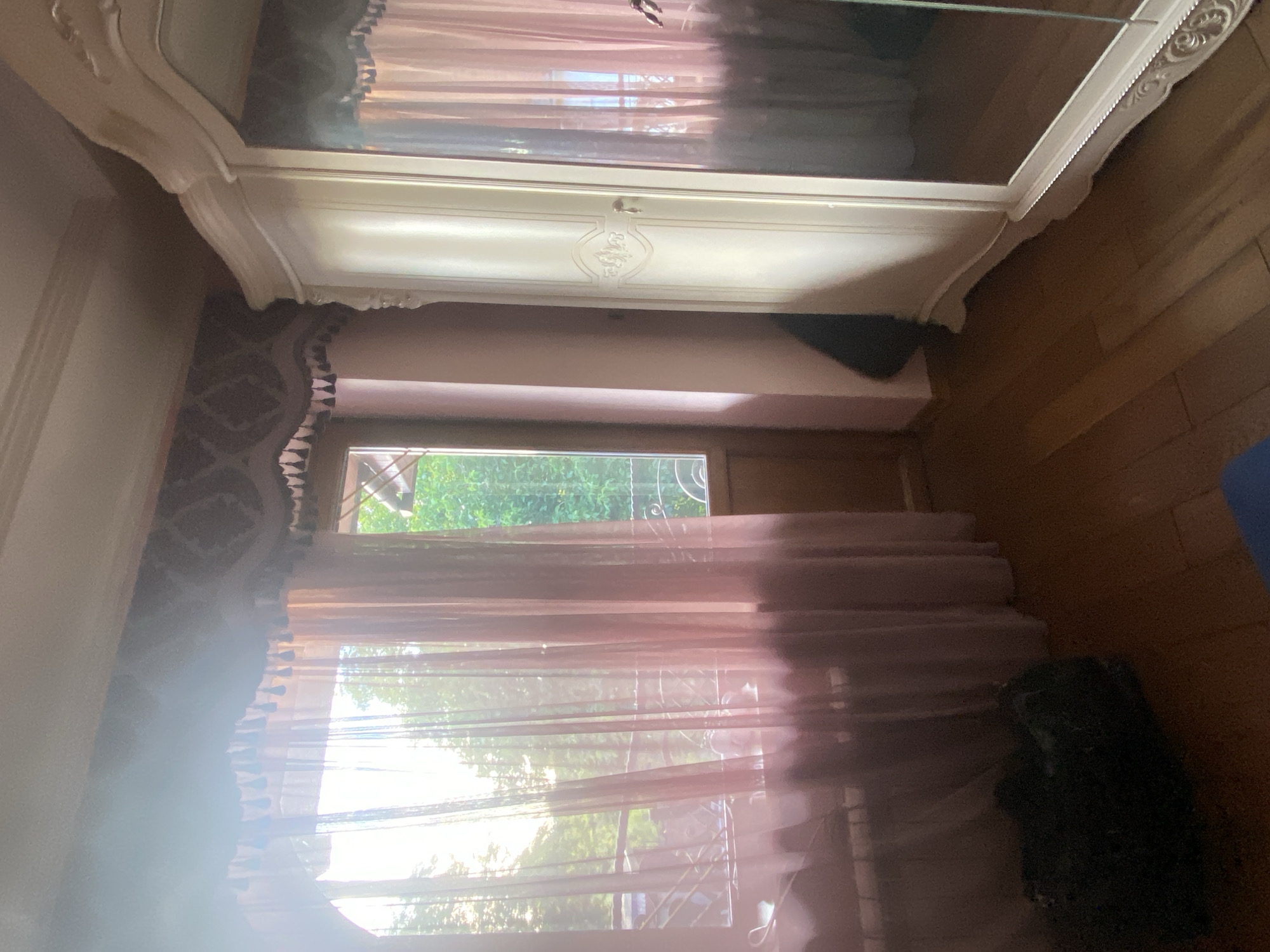 This image depicts an interior room in the city of Unknown, Unknown, illuminated by bright natural light streaming through a large window on the left. The window is adorned with sheer, light pink curtains that are mostly drawn, obscuring a full view of the outside but allowing light to penetrate. Above the window is a decorative valance with dark tassels and a patterned border on the wall. To the right of the main window, a partial view of an open or closed door leading outside is visible, framed by a narrow wooden section and revealing more green foliage. A metal railing is visible beyond the window and door. Below the window, a white radiator is present.
On the right side of the room stands a tall, ornate, cream-colored wardrobe or cabinet. The wardrobe features decorative carvings on its top and bottom edges, with one visible door having an embossed design and a small handle. The right door of the wardrobe is mirrored, reflecting a distorted view of the pink curtains and the bright window. The walls are a light peach or pink color, with a darker patterned wallpaper band above the window. The floor is covered with light brown wooden planks. In the bottom left corner, a dark, possibly green, object or bag rests on the floor, next to a small blue object at the very bottom edge of the frame. No people are present in the scene, and no actions or interactions are depicted.