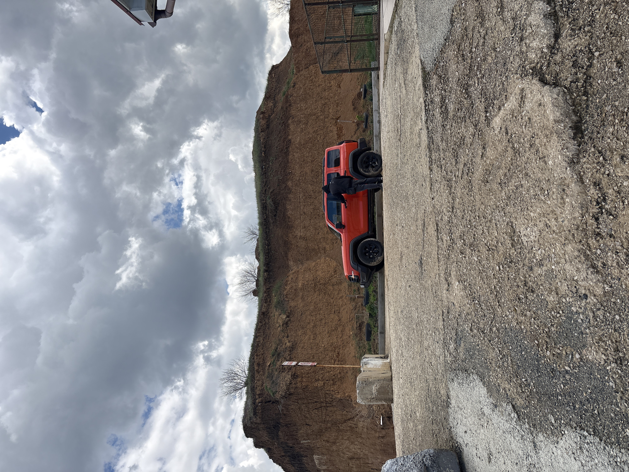 The image depicts an orange Jeep parked on a concrete surface near the edge of a cliff or steep embankment. A person wearing dark clothing is standing next to the driver's side door. The cliff is composed of earth and has sparse vegetation. The sky is overcast with grey clouds, and there are glimpses of blue sky. In the background, there is a metal fence or enclosure. The location is likely near Николаевское сельское поселение, Ukraine, due to the user's prompt. The image is oriented vertically.