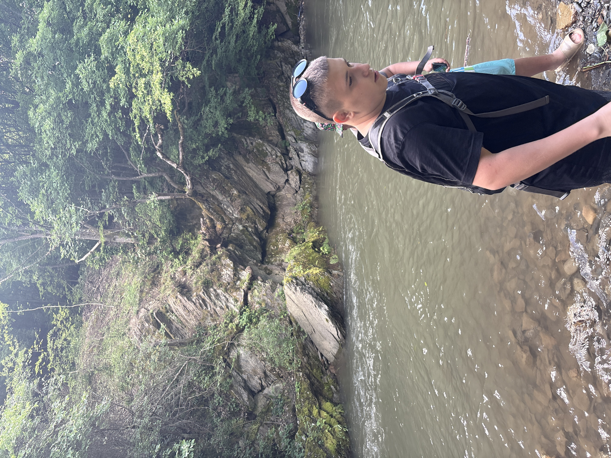 A young male, wearing a black t-shirt, a backpack, and sunglasses perched on his head, stands in shallow water by the bank of a river. His skin tone is fair, and he has short, light-colored hair. Behind him, another person, partially visible, wears a patterned hat and a backpack. The river flows gently, with rocks visible beneath the surface and along the shore. The background features a steep, rocky incline covered with dense green foliage and trees, characteristic of a natural, forested environment. The lighting suggests an overcast day. This scene appears to be in a natural setting, possibly a hiking trail or a recreational area near Lukovo, Bulgaria, given the typical landscape.