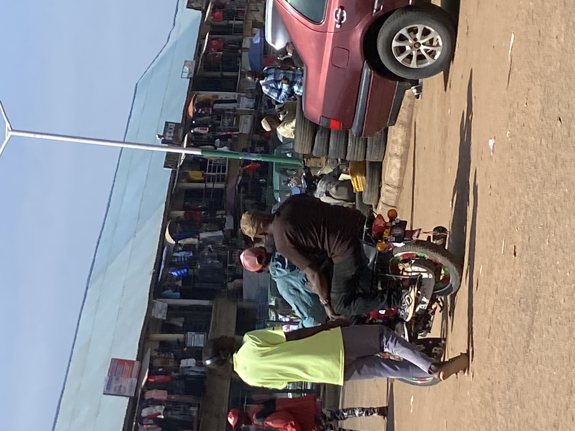 The image shows a street scene in Bukuru, Nigeria, with a focus on a motorcycle carrying three people and a maroon sedan. The motorcycle is in the foreground, with two individuals seated on it and one standing to the left, facing away from the camera. The seated passengers are wearing casual clothing and hats. The motorcycle has colorful decorative elements on its wheels. To the right of the motorcycle is a maroon sedan, partially visible, with its rear wheel in the frame. The background reveals a row of market stalls with goods displayed outside, indicating a busy commercial area. The overall environment is a dusty, outdoor marketplace under a clear sky.