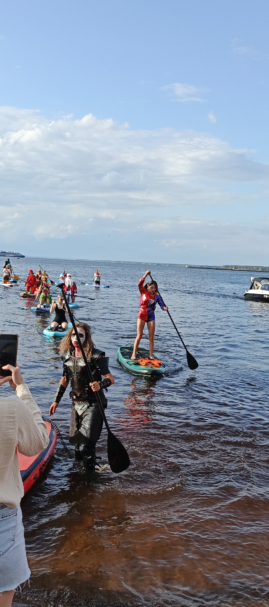 A group of people in costumes are paddleboarding on a body of water, likely a river or lake, under a partly cloudy sky. In the foreground, a person dressed in a dark, armored costume with face paint is standing on a paddleboard and holding a paddle. To their right, another person in a red, blue, and yellow superhero-like costume is also paddleboarding, holding a paddle. Further in the background, a procession of paddleboarders in various costumes, including a Santa Claus costume and red outfits, are visible. A large ship or ferry can be seen in the distance on the left side of the frame, and a small boat is on the right. The water is choppy, and the shoreline with trees is visible in the far distance. The event appears to be taking place in or near the city of Chaikovsky, Russia, possibly along the Kama River, as indicated by the presence of a large body of water and the characteristic landscape.