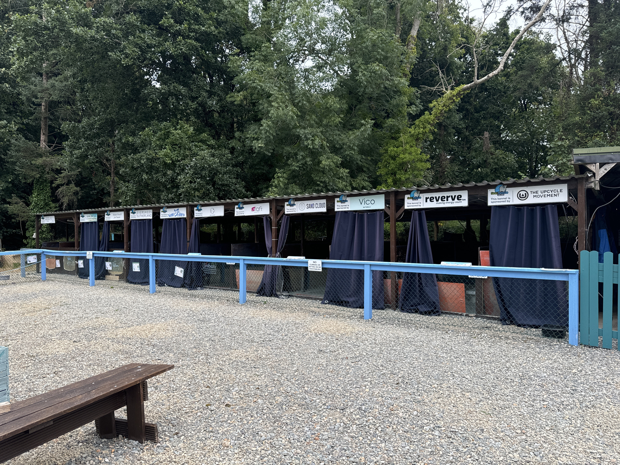 The image depicts an outdoor area with a row of individual kennels, each covered by dark blue curtains. The kennels are situated behind a blue metal fence, which is itself bordered by a gravel-covered ground. In the foreground, a wooden bench is positioned on the gravel. The background is dominated by dense green trees. Above the kennels, a wooden structure supports signs indicating sponsors, including "Eversheds Sutherland," "Now Coco," and "The Upcycle Movement." A small green fence is visible to the right of the main blue fence. The scene appears to be a functional outdoor facility, possibly a dog kennel or a similar animal enclosure, located in Courtown, Ireland. There are no people visible in the image.