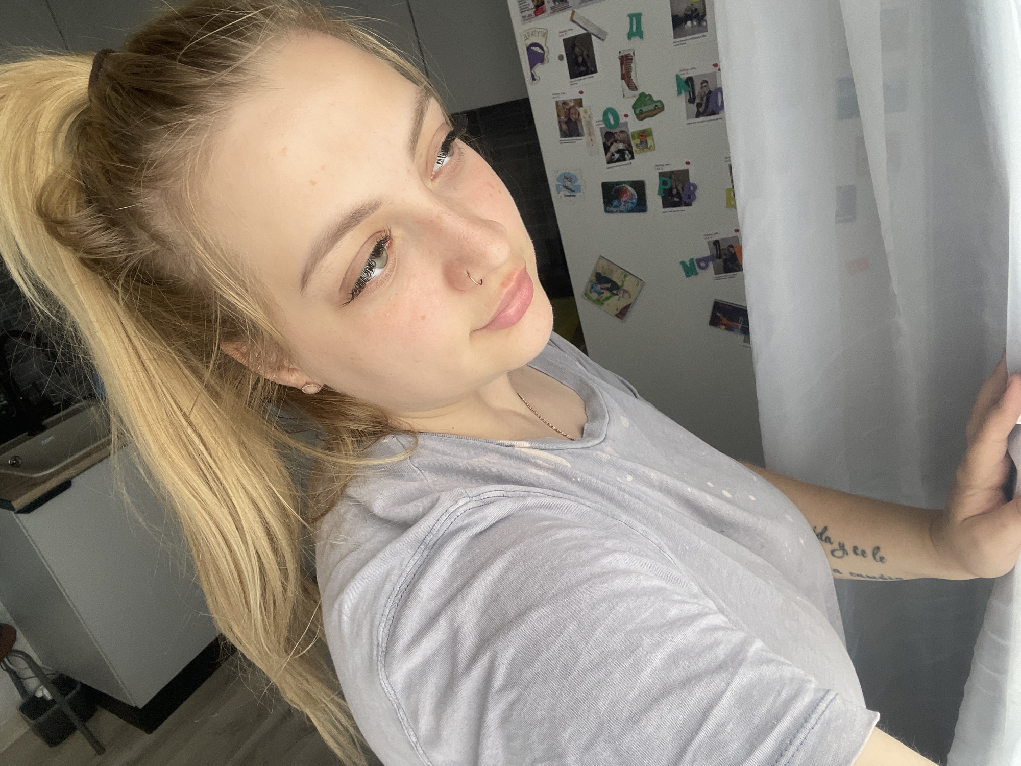 A young, light-skinned woman is shown in a close-up, selfie-style shot, likely indoors. She has blonde hair pulled back in a ponytail, a nose ring, and fair skin with freckles. She is wearing a light blue or grey t-shirt. In the background is a refrigerator covered with magnets and photos. White curtains are partially visible on the right side of the frame. A sink is visible in the lower left. There is a tattoo on her inner forearm. The environment appears to be a kitchen. There is nothing to specifically indicate Izhevsk, Russia.