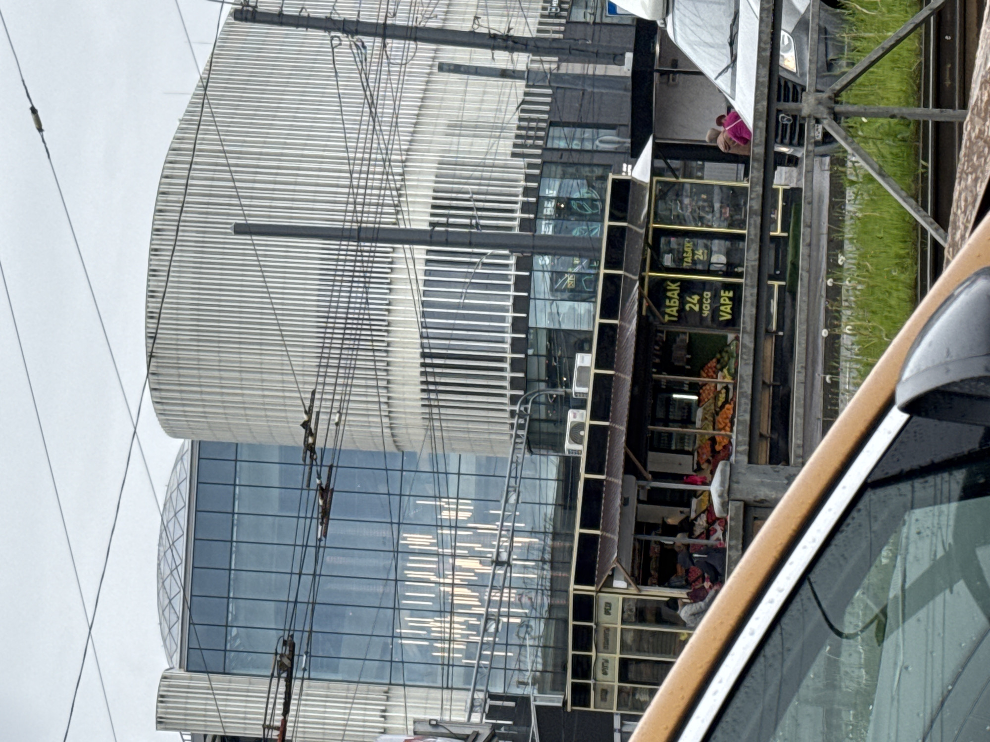 The image shows a street scene in Saint Petersburg, Russia. A portion of a car's windshield and frame is visible on the right. In the background is a modern building with a curved, striped facade. There is a structure, possibly a tram or railway line, with vegetation growing alongside it. A small retail establishment advertising "TABAK 24 часа VAPE" (Tobacco 24 hours Vape) is present. Several people are seen near the retail establishment, some of whom appear to be interacting with the store. Overhead, electrical wires and support structures are visible against a cloudy sky.