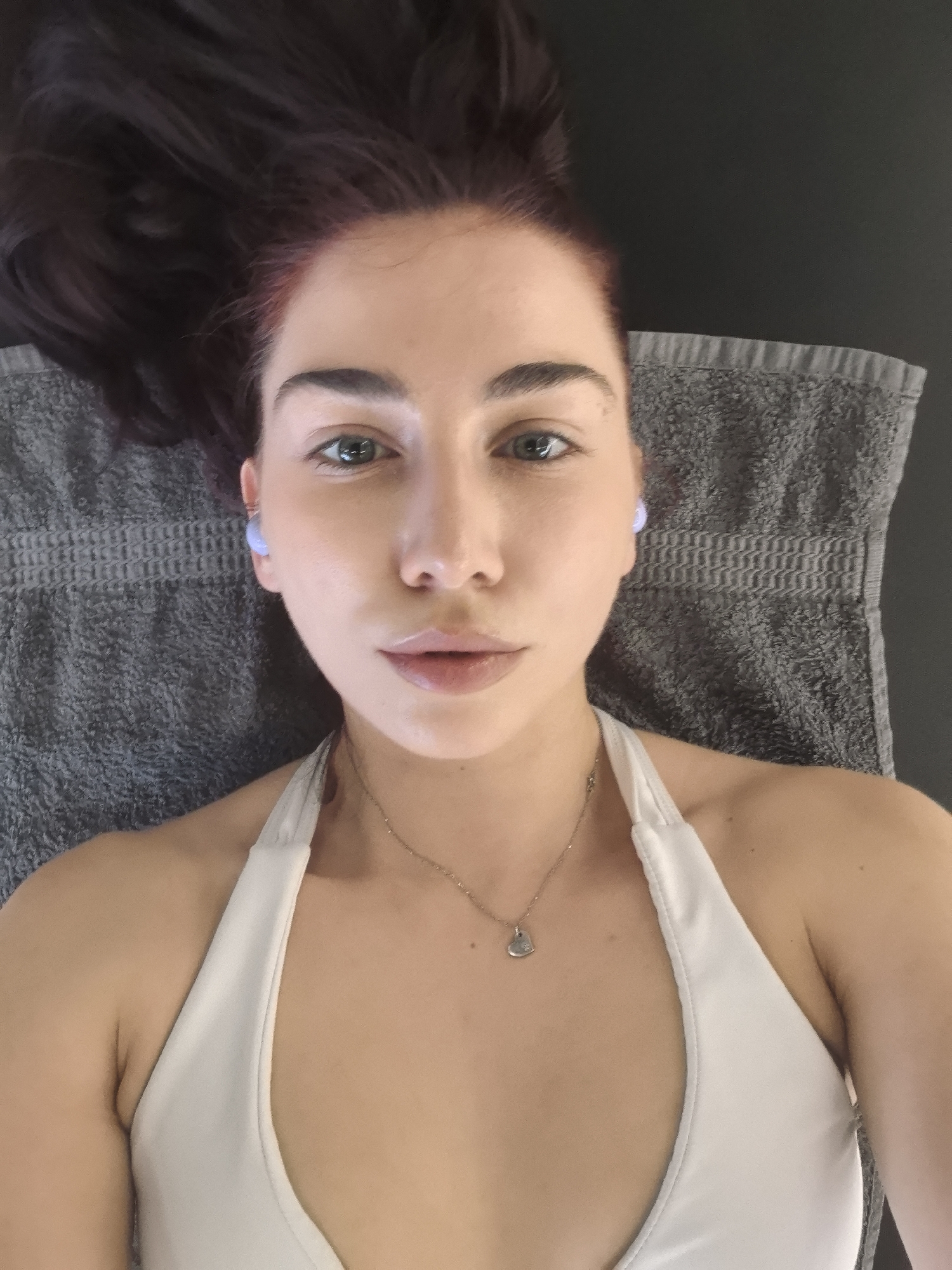 A woman is lying on a gray towel, wearing a white tank top and a silver necklace. She has dark hair with red undertones, green eyes, and appears to be wearing earbuds. The location is unknown.
