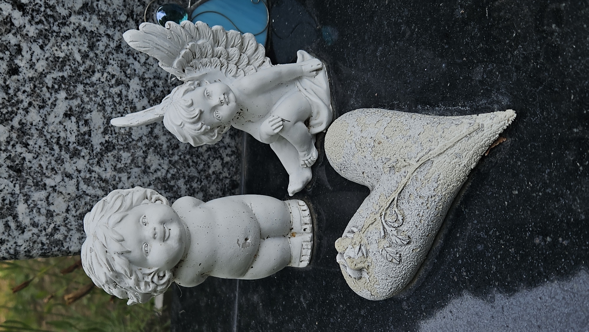 Three white decorative figures, likely made of plaster or ceramic, are arranged on a dark, polished surface, possibly a gravestone. In the foreground, a heart-shaped object with a raised floral motif rests on its side, its surface weathered and textured. Behind the heart, a plump cherubic figure stands with its hands clasped behind its back. To the right, a winged cherub sits with one leg bent and the other extended, looking towards the viewer. A dark grey granite slab with visible speckles serves as the backdrop. The scene appears to be outdoors, with blurred greenery visible in the upper left corner, suggesting a garden or cemetery setting. A hint of a blue decorative element is visible on the far right. The image has a somber, reflective quality.