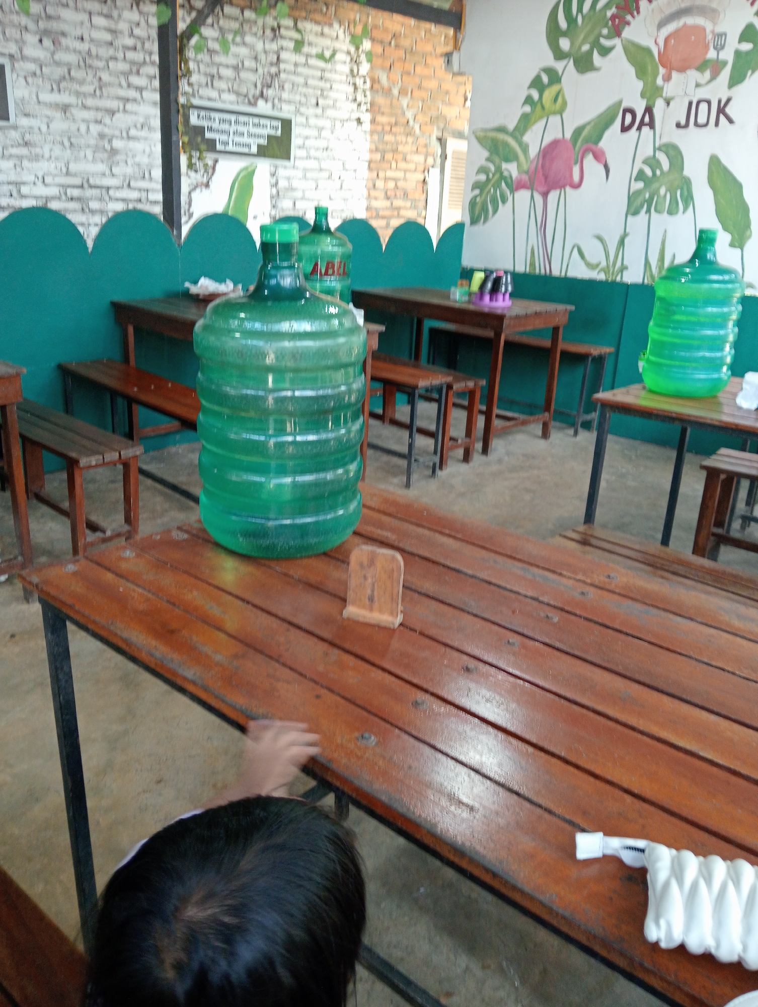 The image depicts an outdoor seating area, likely a restaurant or cafe, in Campago, Indonesia. Wooden tables with black metal legs are arranged around green benches. In the foreground, a large green water jug sits on a wooden table, and a young child's head and hand are visible, reaching towards the table. In the background, another green water jug is on a table, and a mural adorns the wall with tropical plants and a pink flamingo. The overall scene is casual and appears to be an establishment serving drinks.