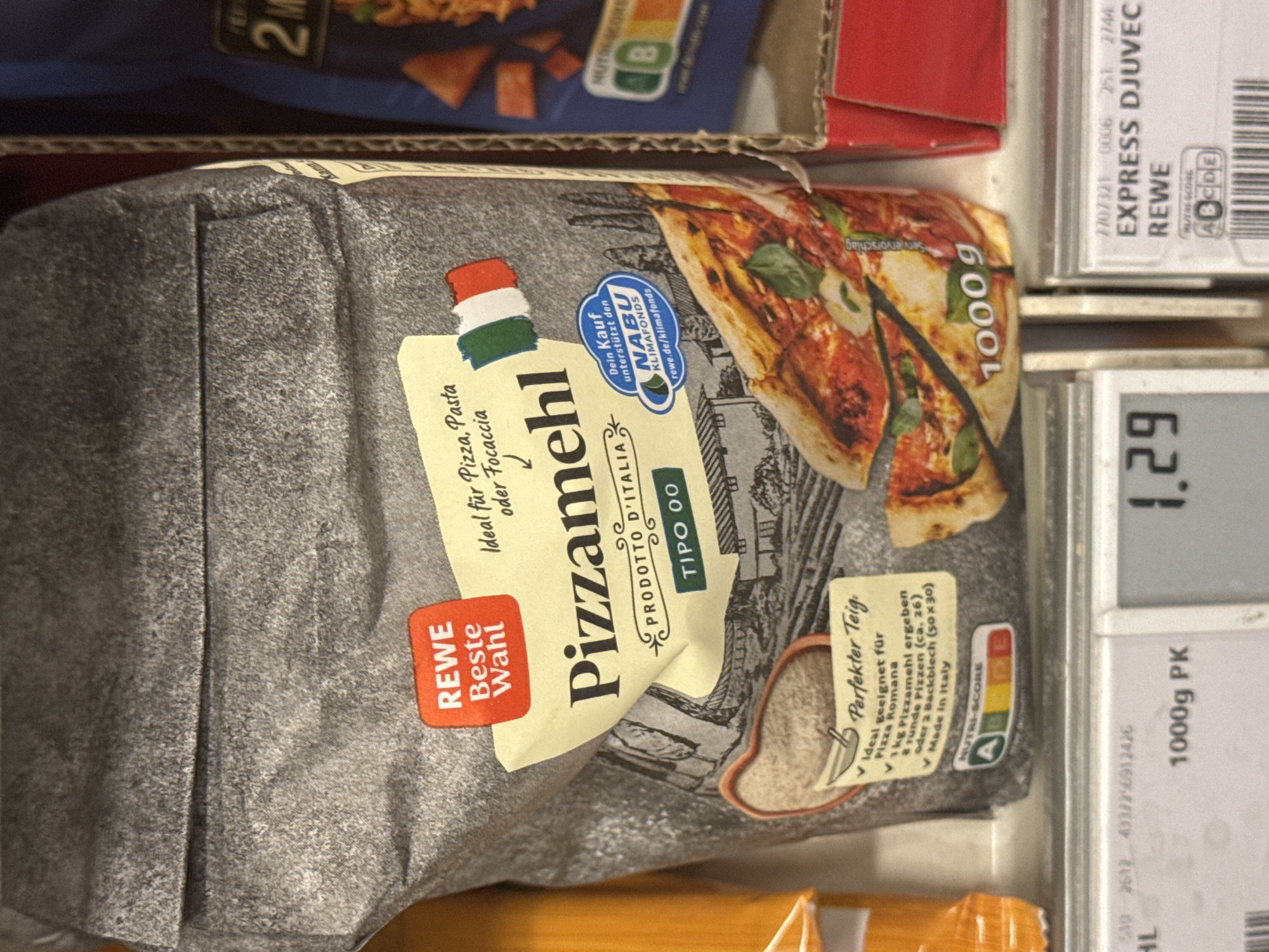 This is a close-up, slightly angled photograph of a bag of "Pizzamehl" (pizza flour) by REWE. The bag is predominantly grey with a large, vibrant image of a pizza on the right side, featuring melted cheese and basil. The flour bag is lying on a shelf, with other products visible above and below.
To the left of the flour bag, there is a red and white label that says "REWE Beste Wahl," indicating it's a house brand. The flour itself is described as "Ideal für Pizza, Pasta oder Focaccia" (Ideal for pizza, pasta, or focaccia) and is specifically for "Pizza Romana." The bag also prominently displays "PRODOTTO D'ITALIA" and "TIPO 00," signifying its Italian origin and type of flour.
A green and blue circular logo for "NABU KLIMAFONDS" is visible, suggesting that purchasing this product supports a climate fund. A "NUTRI-SCORE" rating of "A" is shown, with a scale indicating it is a healthy choice. The bag contains "1000g" of flour, and a serving suggestion indicates that 1kg of this flour yields approximately 8 round pizzas or 2 baking sheets.
To the right of the flour bag, a white package with black text is visible, featuring "EXPRESS DJUVEC REWE." A "NUTRI-SCORE" of "B" is also displayed on this item. Below the flour bag, a digital scale displays "1.29," and another package shows "1000g PK."
The setting appears to be a retail environment, likely a grocery store or supermarket shelf in Neubrandenburg, Germany, given the context. The lighting is bright, typical of an indoor retail setting, and there are no indications of weather or time of day. The overall impression is one of a product display, showcasing a food item with clear labeling and nutritional information.