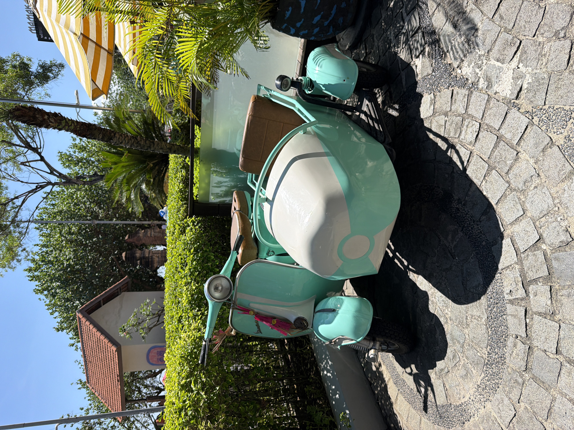 The image shows a light blue vintage Vespa scooter with a sidecar parked on a cobblestone surface. The sidecar is white with a light blue stripe. The cobblestones are arranged in a circular pattern. Behind the scooter is a tall green hedge. In the background, there's a building with a brown tiled roof and trees. A yellow and white striped awning is visible in the upper left corner. The photo appears to be taken outdoors on a sunny day. The location is likely Kuta, Indonesia, considering the provided information.
