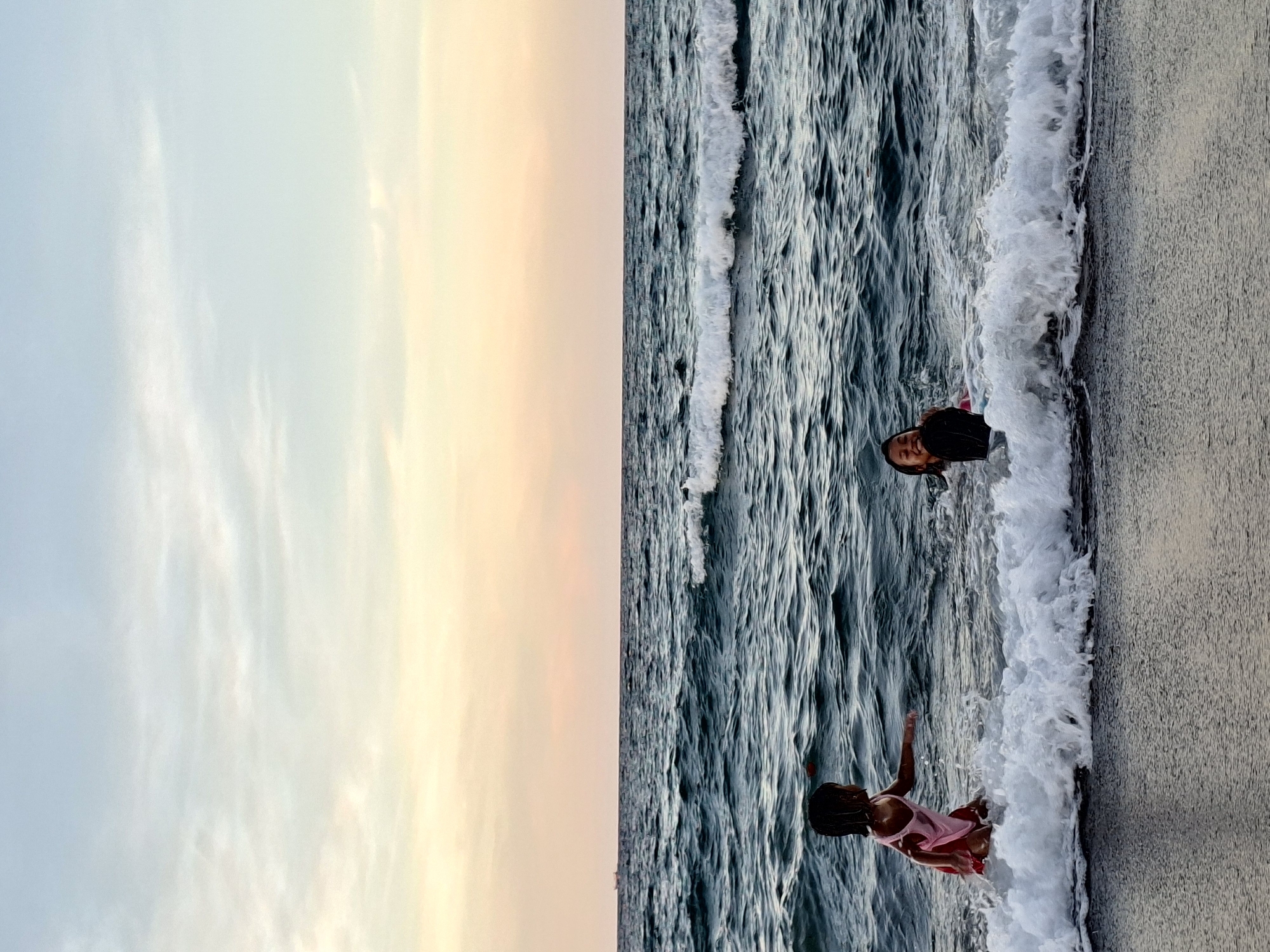 The image depicts a beach scene at sunset. The sky is divided vertically, with a pale blue section on the left transitioning to shades of orange and yellow on the right. A body of water with choppy waves occupies the middle ground. Two people are in the water near the shoreline. One person, wearing a black shirt, is further out in the water. The other, wearing a pink dress, is closer to the shore, with the waves lapping around her. The beach itself is visible on the right edge of the image. Based on the prompt, the location is Lhokseumawe, Indonesia.