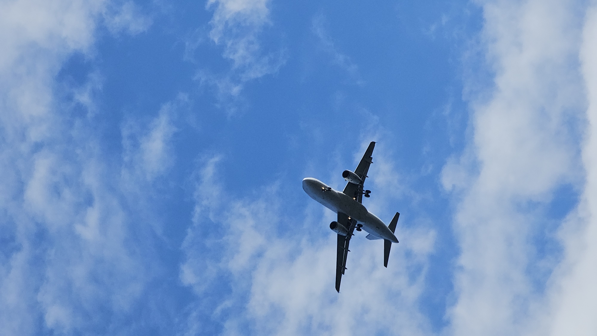 A commercial airplane is flying in a blue sky with scattered clouds. The airplane is flying low in the sky, likely landing or taking off. It's difficult to ascertain the exact location within Varna, Bulgaria from this image alone, but the view suggests an area with open sky, such as near the airport, or a coastal area. The plane is viewed from below. Landing gear is deployed.