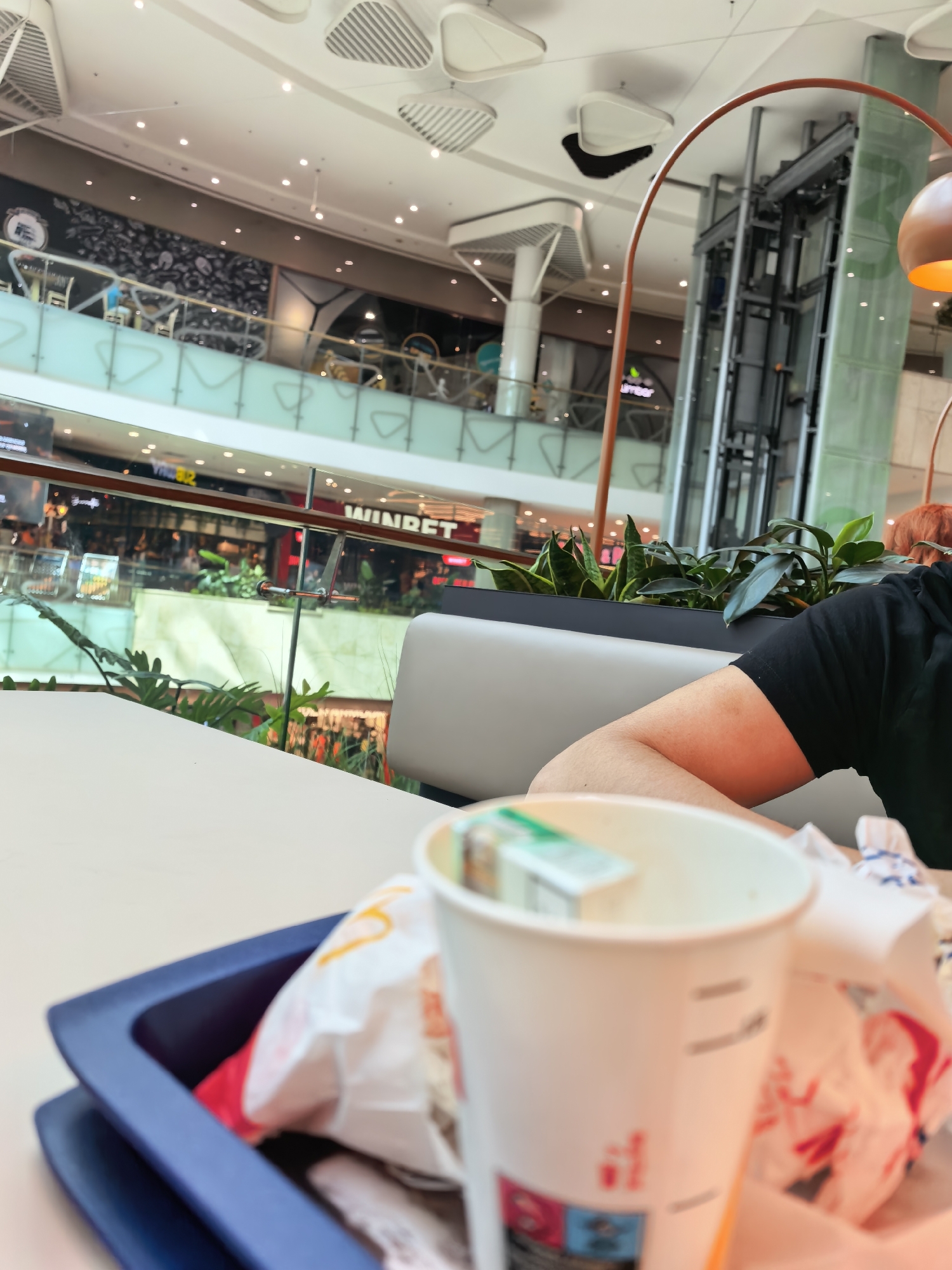 The image shows a table in a mall food court, with a blue tray containing fast food packaging and a cup. On the table, there's a white paper cup with a pack of cigarettes inside. A person's arm and shoulder are visible in the lower right corner, wearing a black shirt. The background features the mall's interior with multiple levels, plant decorations, and a glass elevator. A sign with "WINBET" is visible in the background. The location is in the city of Sofia, Bulgaria.