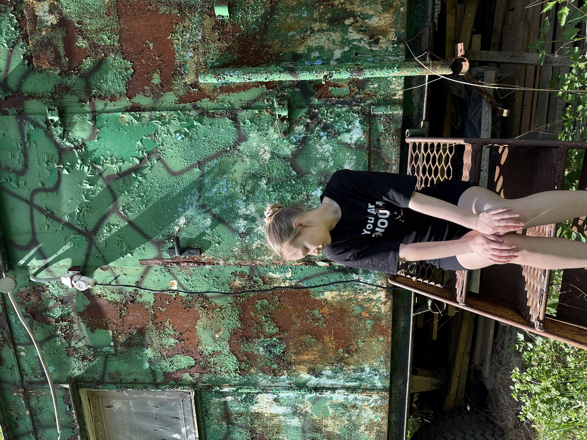 A young woman with fair skin and blonde hair pulled back in a bun sits on the rusty metal steps of what appears to be an abandoned, green, metal structure. She is wearing a black t-shirt and black shorts, and her hands are resting on her knees. The metal structure shows significant rust and peeling paint, revealing layers of green and brown. An electrical conduit is visible on the structure. The area around the structure is overgrown with weeds and some scattered wooden debris. The location is not possible to determine based only on this image.