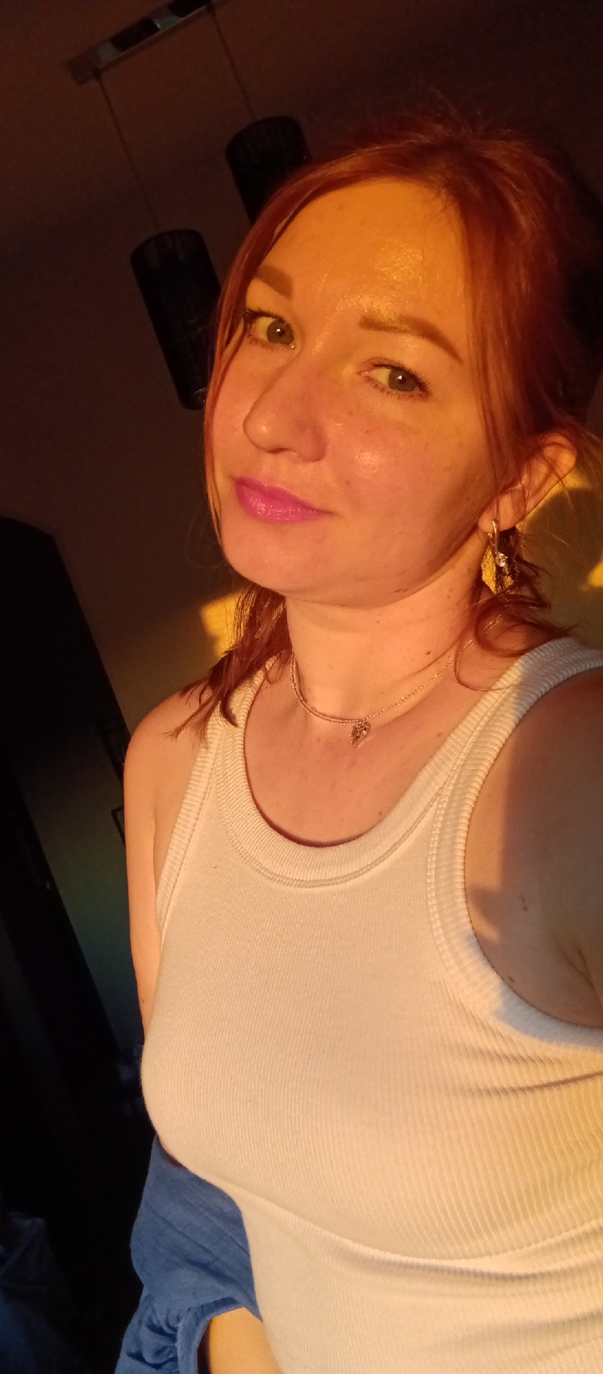 A medium shot captures a woman with fair skin, reddish-brown hair, and pink lipstick. She is wearing a white tank top and a delicate necklace, and her left ear is adorned with a gold earring. The lighting in the image is warm, casting a yellowish glow on her face and body, with shadows on the left side of the frame. In the background, there is a dark object resembling a lamp or part of a lighting fixture. A blue garment is partially visible near her torso. The specific location is unknown.