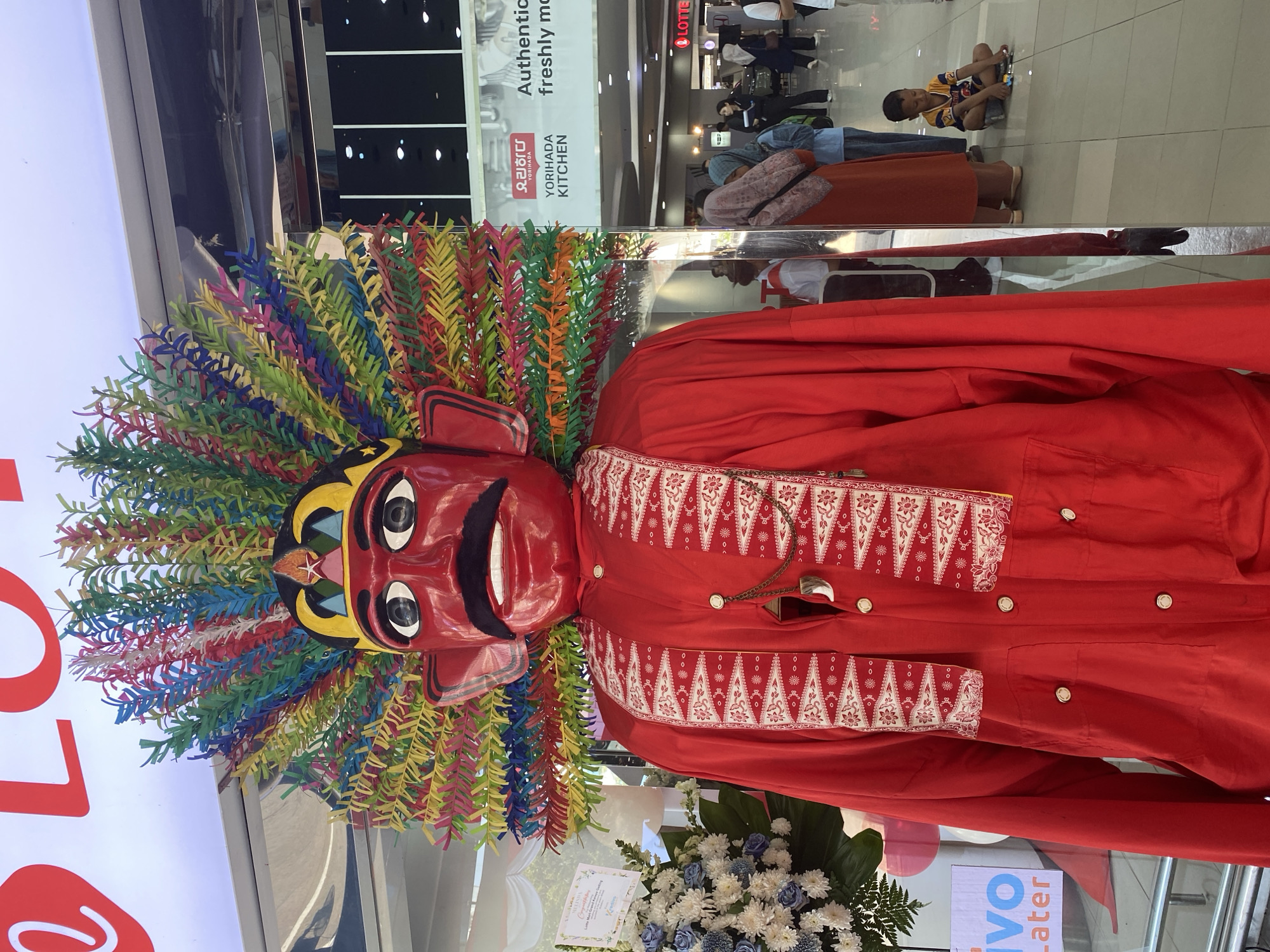 The image shows a large, red effigy of a figure, likely a traditional Indonesian Ondel-ondel, standing in what appears to be an indoor public space. The effigy has a red face with large black eyes, a black mustache, and a prominent red mouth. It is adorned with a colorful headdress made of paper strips in various hues of red, yellow, blue, green, and pink, resembling feathers. The effigy is wearing a bright red tunic with white and red patterned trim on the chest and a gold-colored chain with a tooth-like pendant.
In the background, there is signage for "YORIHADA KITCHEN" and "LOTTE," suggesting a commercial area, possibly a mall or shopping center in North Jakarta, Indonesia. Several people are visible in the background, including a woman in an orange hijab, a woman in a blue hijab, and a young boy sitting on the floor playing with toy cars. There are also flower arrangements to the left of the effigy. The lighting suggests an indoor environment with overhead lights.