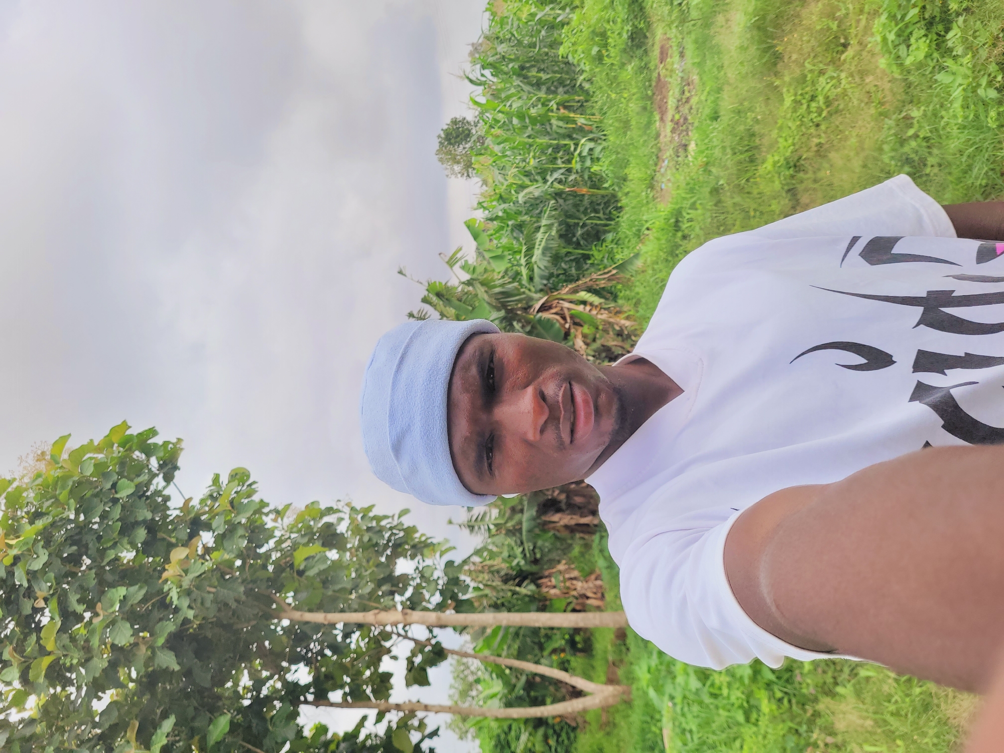 The image, taken in Bukuru, Nigeria, features a dark-skinned male subject, appearing to be taking a selfie, looking directly at the camera with a neutral to slightly smiling expression. He is wearing a light blue head covering, possibly a durag or skullcap, and a white short-sleeved t-shirt with a prominent graphic design on the right side, composed of black stylized shapes and potentially hints of pink. His right arm and shoulder are visible in the foreground, indicating the self-portrait style.
The setting is outdoors, characterized by abundant green vegetation suggesting a rural or agricultural environment. To the subject's left, there are trees with dense green foliage. Further into the background, particularly to the right, there is a wide expanse of tall, lush green plants, resembling corn or other field crops, along with broader leaves possibly from banana trees. Patches of lighter green ground are also visible. The sky, seen in the upper left, is light and overcast, indicating daytime conditions with diffused light, suggesting a cloudy day without direct sunlight. There is no clearly legible text on the t-shirt graphic.