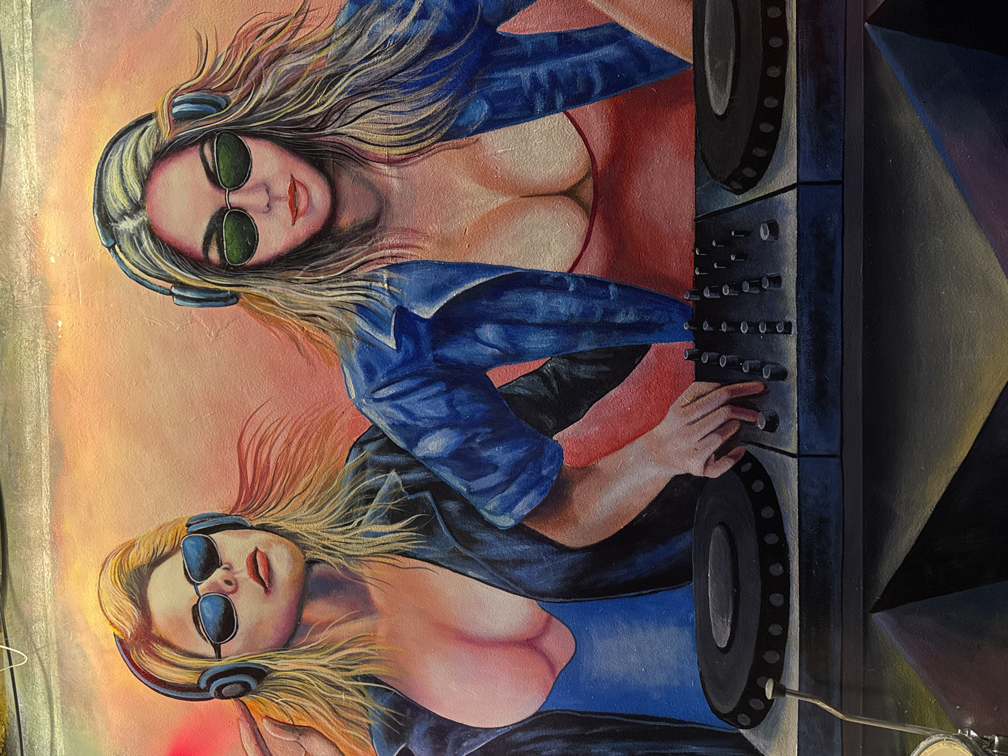 The image shows a mural depicting two women dressed as DJs. Both have blonde hair, wear sunglasses and headphones, and are positioned behind DJ equipment. One woman wears a blue jacket over a red top, while the other wears a blue top. The mural features a pink and orange background. The image was taken in Đà Nẵng, Vietnam.
