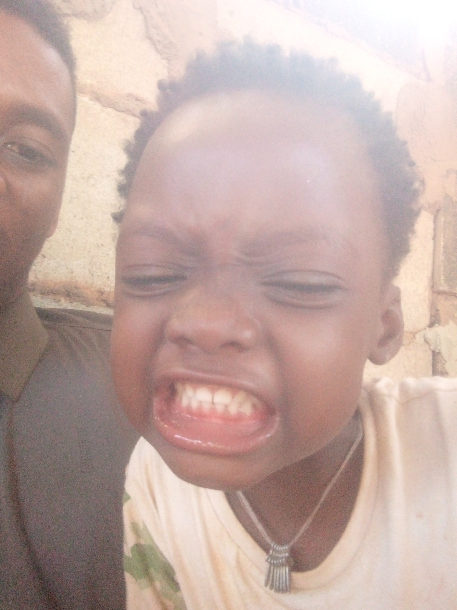 The image depicts a close-up of two individuals. On the left, a portion of an adult's face is visible, with dark skin and a closed eye. They are wearing a brown collared shirt. To the right, a child with dark skin and curly black hair is making a face, eyes closed, and teeth bared. They are wearing a light-colored t-shirt and a silver necklace with a pendant. The background appears to be an exterior wall made of earth-colored bricks. The location is specified as Unknown, Unknown.