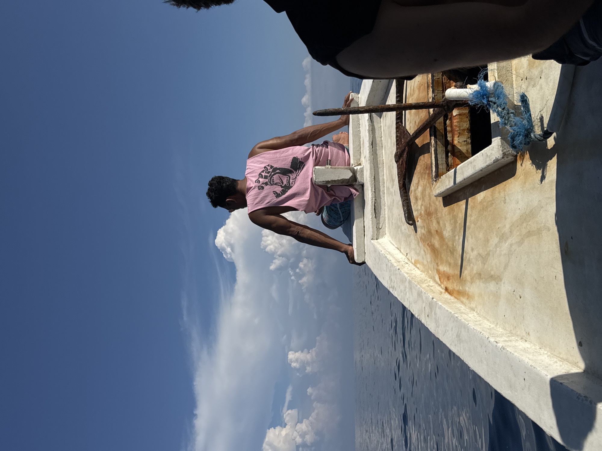 A man wearing a pink tank top with a black graphic and blue shorts sits on the edge of a boat. He dangles his feet over the side, his gaze directed downwards. The boat, which is old and weathered, has a rusty anchor. The sea is calm, with a few waves visible. The sky is blue with puffy white clouds. The location is Unknown, Indonesia.