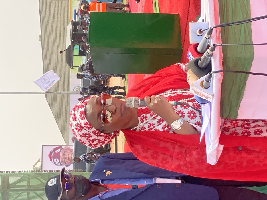 The scene depicts an outdoor public event taking place in Ifira, Nigeria. A woman, the primary subject, stands centrally, holding a grey microphone to her mouth with her right hand, actively speaking. She wears a red and white patterned headwrap, dark sunglasses, a red sheer overlay shawl, and a white and red patterned dress. A multi-strand white bead necklace is visible around her neck, and a silver wristwatch adorns her left wrist. She stands on a red carpet, positioned behind a table draped with white, green, and red fabric. On this table, several news microphones, including one labeled "NTA News," and white papers are arranged.
To the left of the speaking woman, a man is partially visible, wearing a black baseball cap, clear-rimmed glasses, and a black face mask. He has a dark blue jacket with a small badge. In the mid-ground, a green rectangular podium is visible behind the woman's right side. Beyond this, numerous individuals, including several uniformed personnel in dark blue uniforms, some with helmets, are observable. One person in an orange safety vest stands further to the right.
The background features a large billboard displaying the portrait of an older man wearing a white cap and traditional attire. A white flag with a dark logo and text is positioned to the right of the billboard. The area behind the immediate event setup consists of structures, possibly temporary, and open, sandy ground under a bright, somewhat hazy sky.