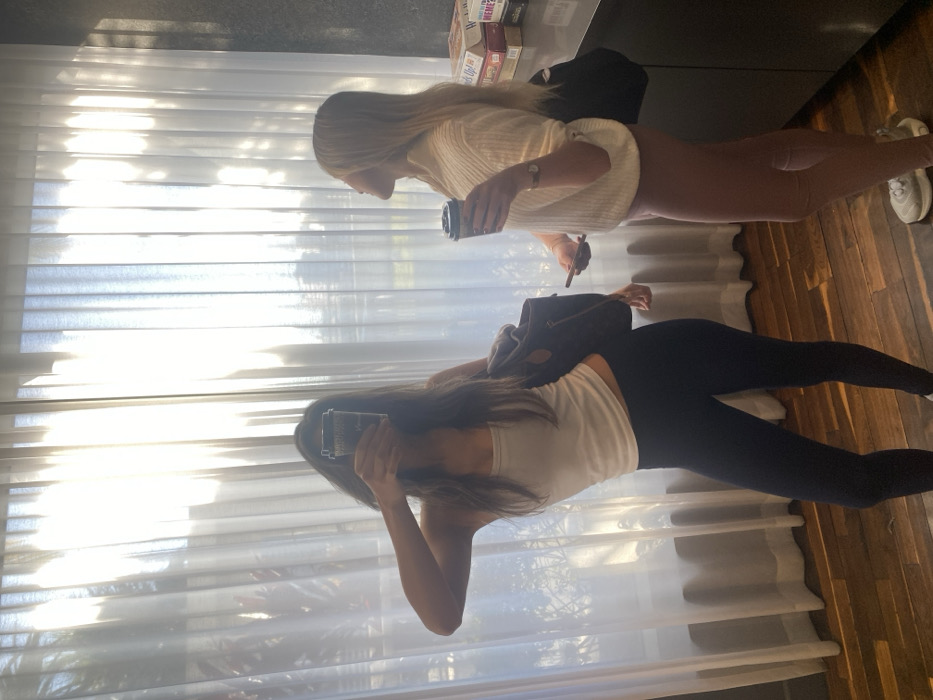 This image depicts two adult females standing within a well-lit contemporary interior space in Miami, United States of America. The floor is constructed from dark wooden planks. Behind the subjects, a large window is covered by sheer white curtains, through which bright sunlight penetrates, illuminating the room and casting shadows, while faintly revealing outdoor vegetation.
On the left, a female with long dark hair, clad in a white sleeveless crop top and black leggings, holds a dark-colored disposable coffee cup directly in front of her face, obscuring her features. A dark brown shoulder bag with light trim is visible on her left side. On the right, a second female with long blonde hair, wearing a cream-colored ribbed long-sleeved sweater, light brown leggings, and white sneakers, stands in profile. She holds a dark-colored disposable coffee cup in her left hand and a smartphone in her right, looking upwards and to her left. A watch is visible on her left wrist.
Behind the second female, a dark-colored counter or cabinet features a stack of board game boxes, including "Heads Up!" and "What Do You Meme?". No direct interaction between the two individuals is observed, though they occupy the same space.